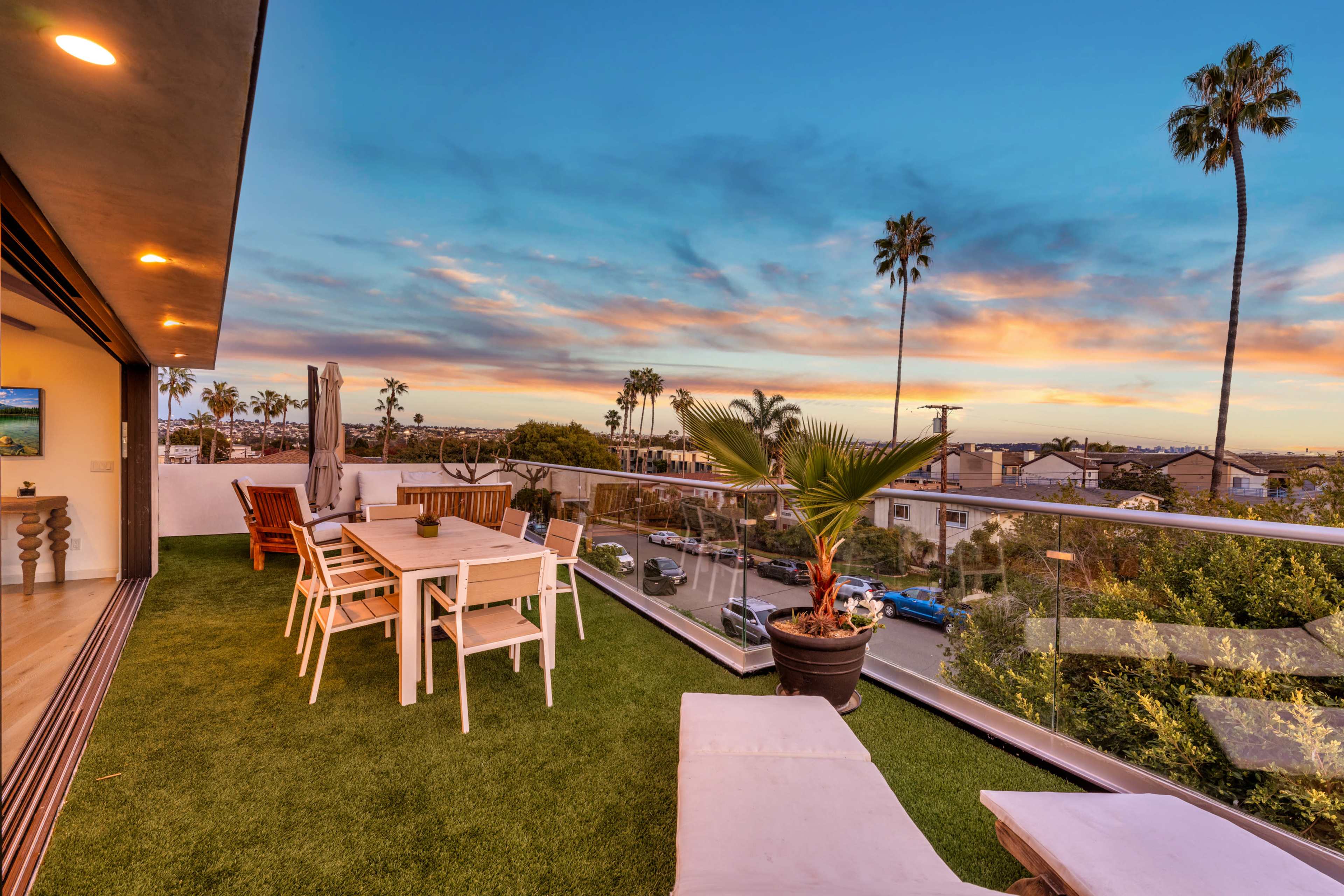 A modern balcony features a dining table, lounge chairs, and potted plants with a view of palm trees and a colorful sunset.