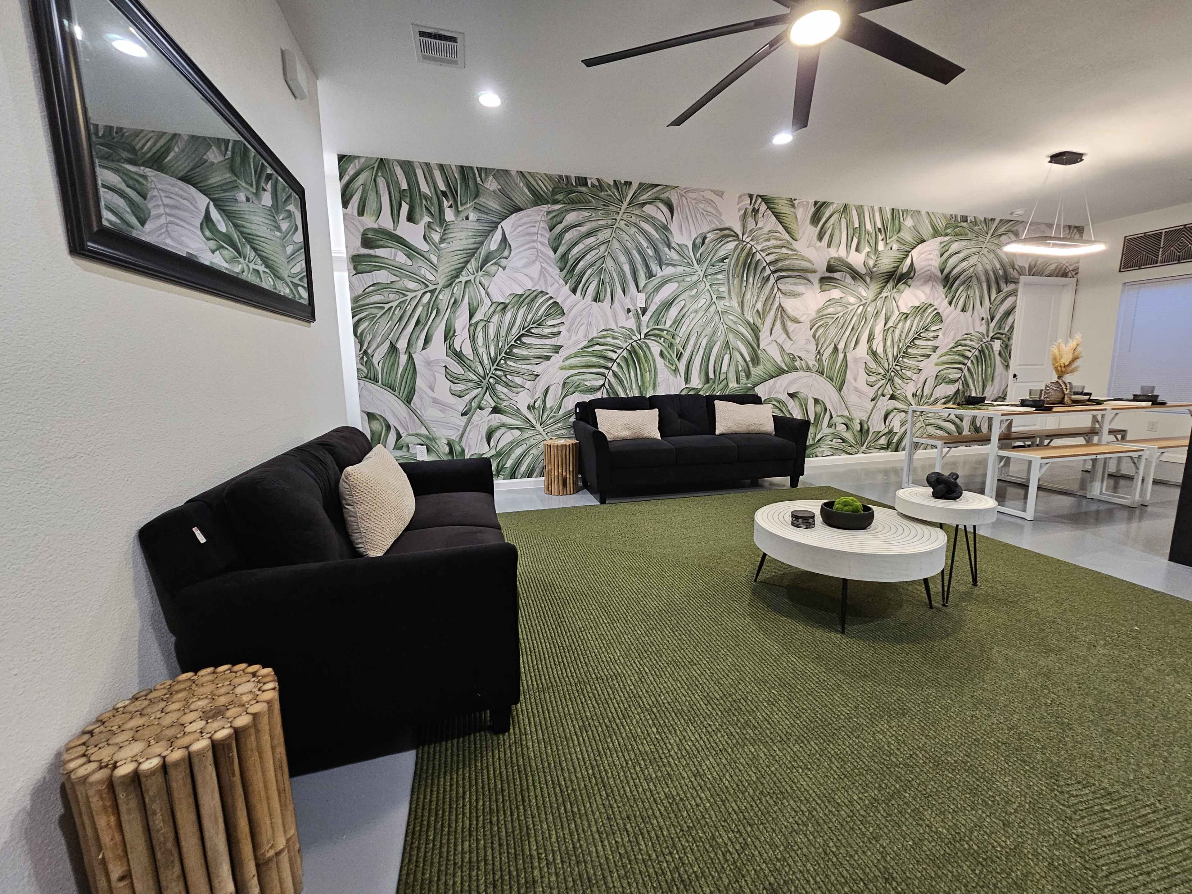 A living room features two black sofas, a round coffee table, and a large tropical leaf wallpaper, complemented by a green area rug.