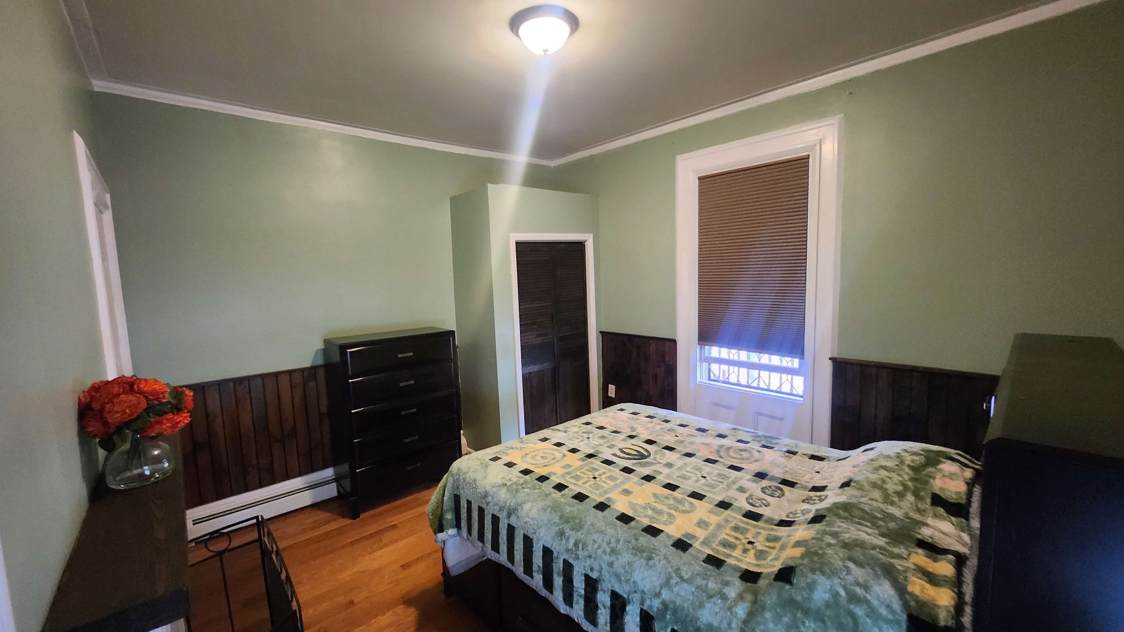 A tidy bedroom with a bed covered in a green and patterned quilt, a wooden dresser, a closet, and a window with blinds.