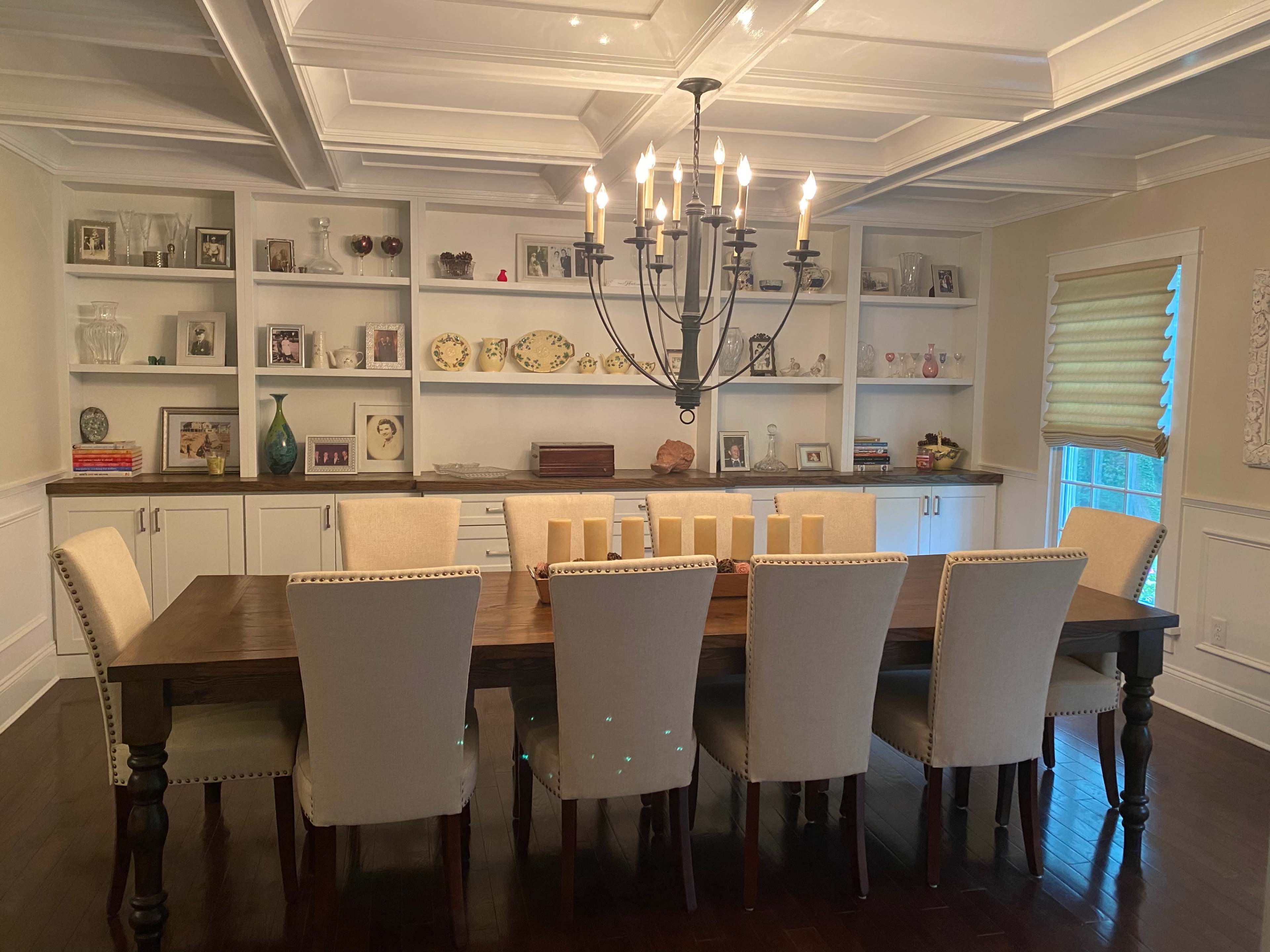 A large dining table with eight upholstered chairs is positioned in a well-lit room featuring built-in shelves adorned with decorative items and family photographs.