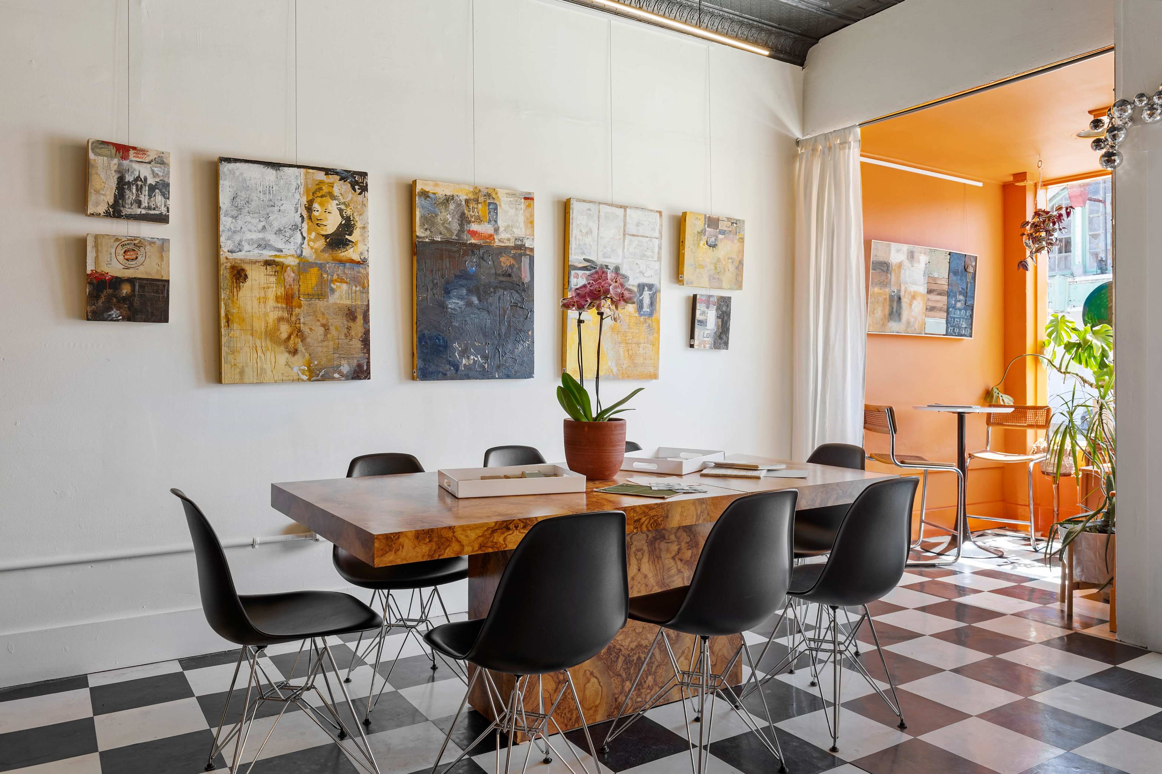 A dining area features a large wooden table surrounded by black chairs, with abstract art displayed on the walls and a plant next to a bright orange nook.