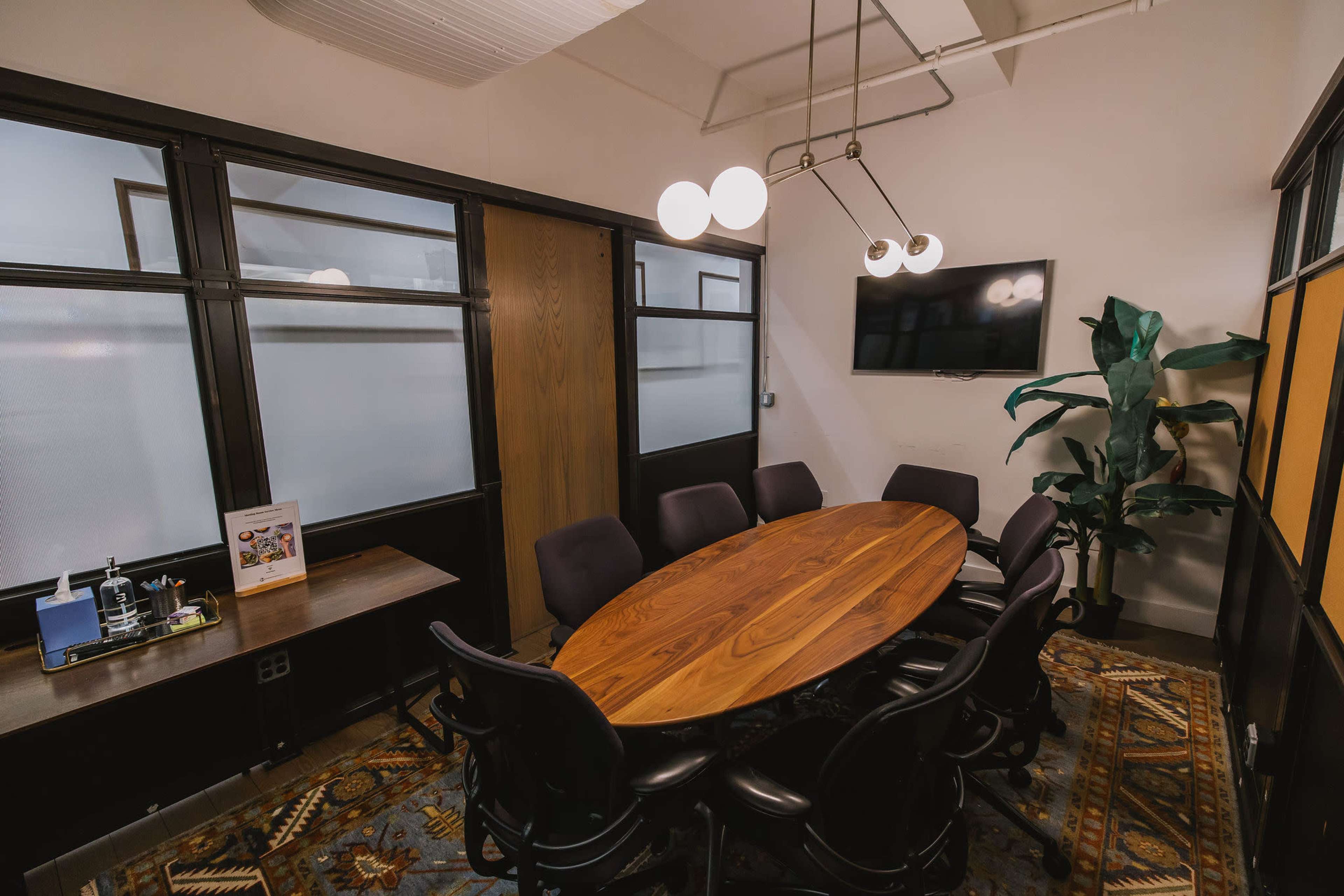 A modern conference room features a large oval wooden table surrounded by black chairs, with a TV mounted on the wall and a plant in the corner.