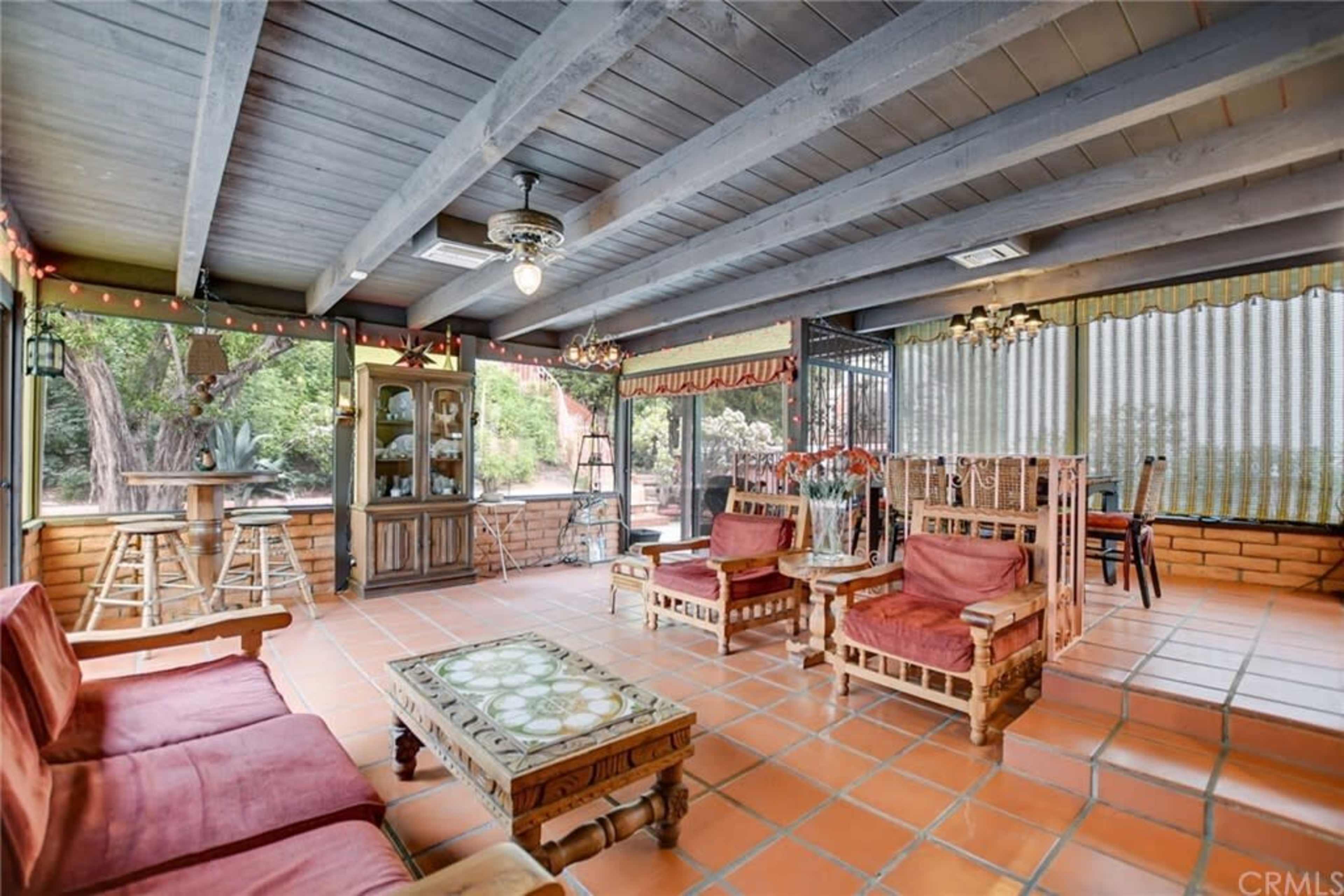 The image shows a spacious sunroom with terracotta tiles, wooden beams, and a mix of seating areas, including chairs and a coffee table.