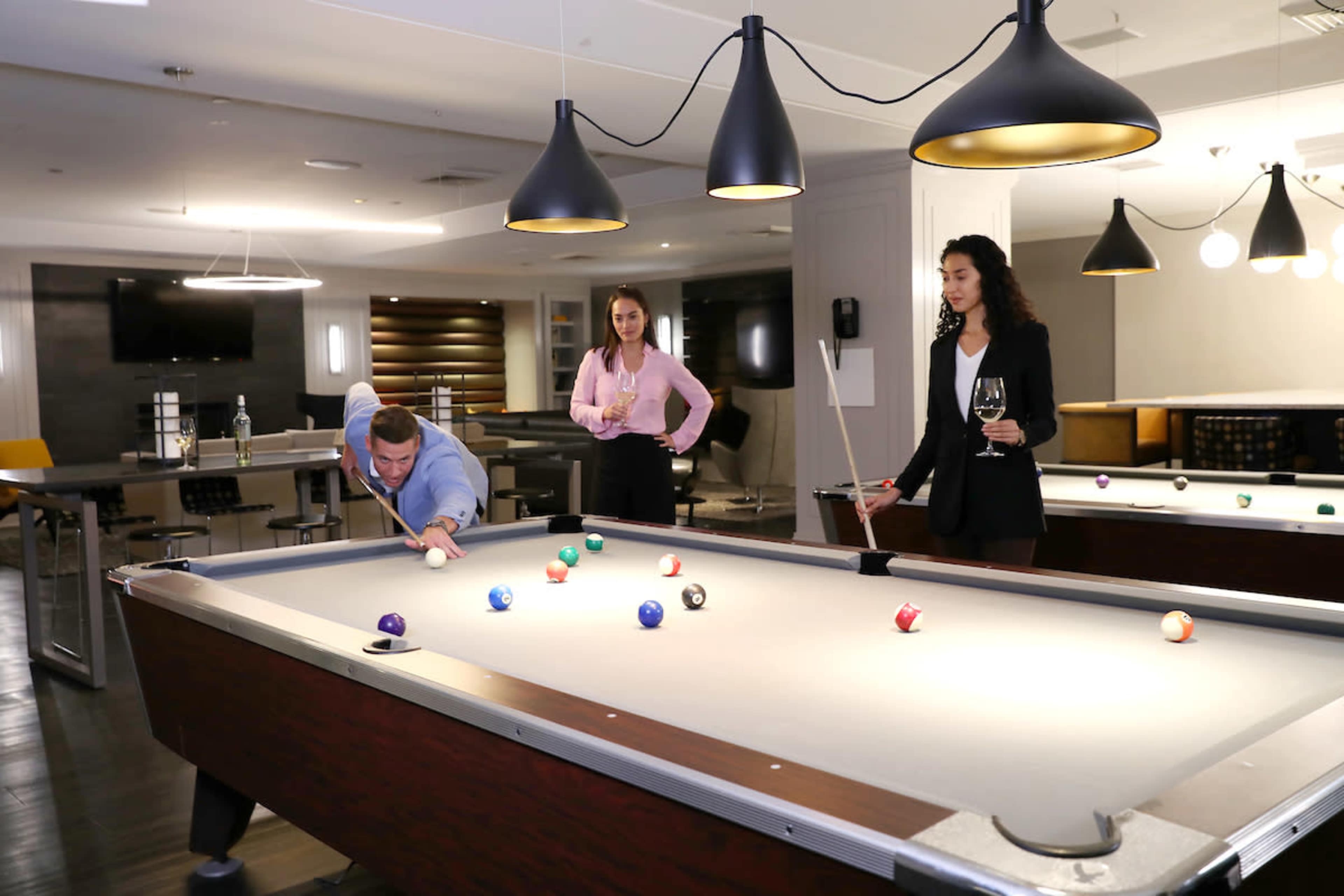 A man leans over a billiards table, preparing to take a shot, while two women stand nearby, one holding a glass of wine.