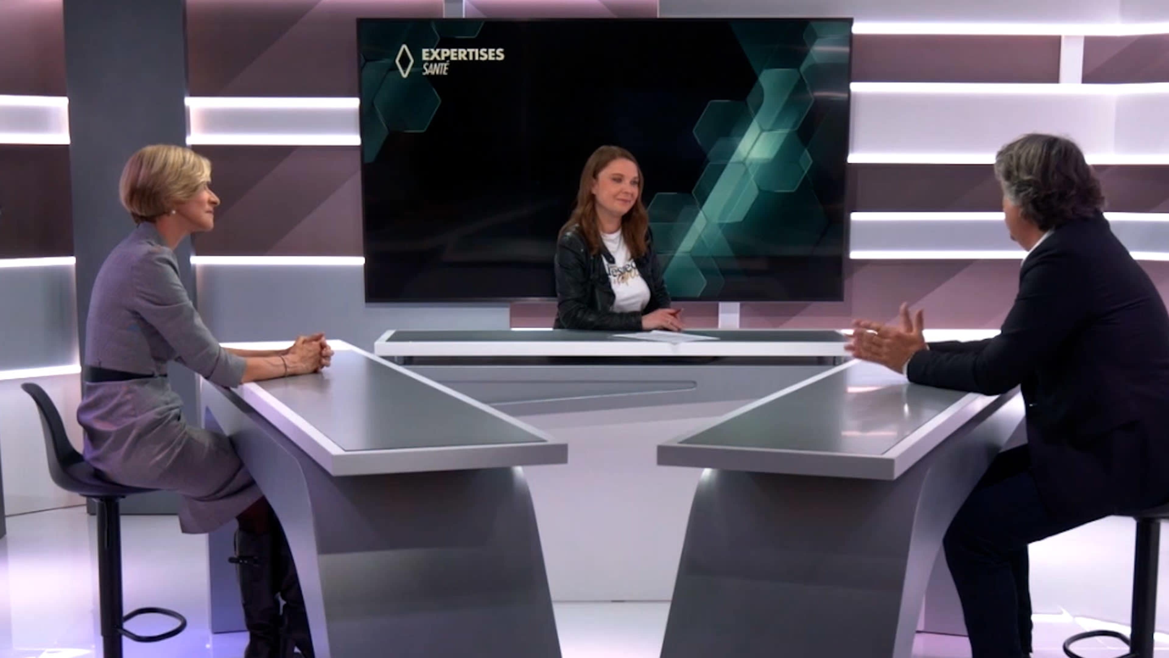Three individuals are seated at a modern studio table, with one person presenting in front of a large screen displaying the word "EXPERTISES."
