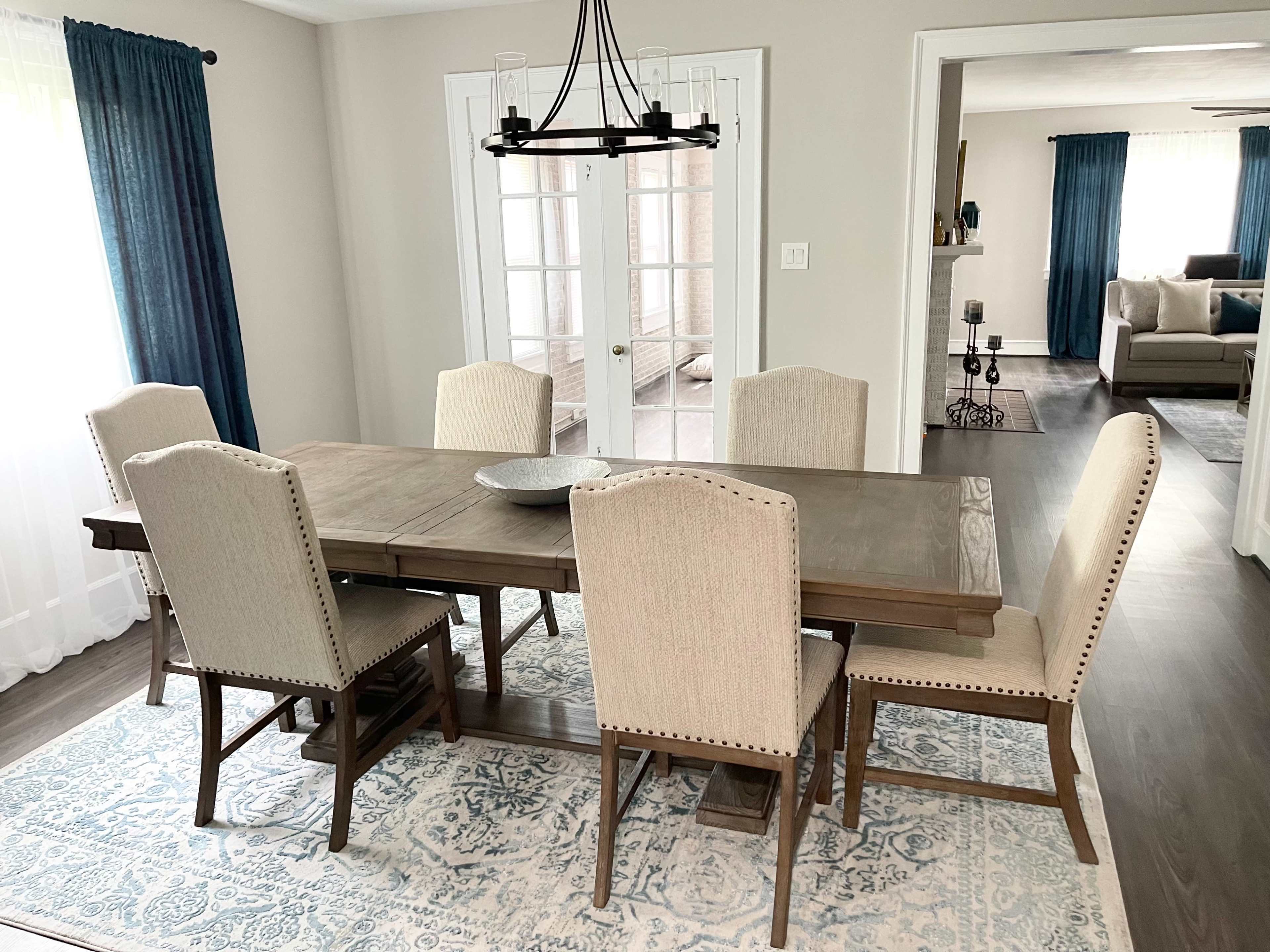A wooden dining table with six upholstered chairs is set on a patterned rug in a well-lit dining room, featuring large windows and a chandelier above the table.