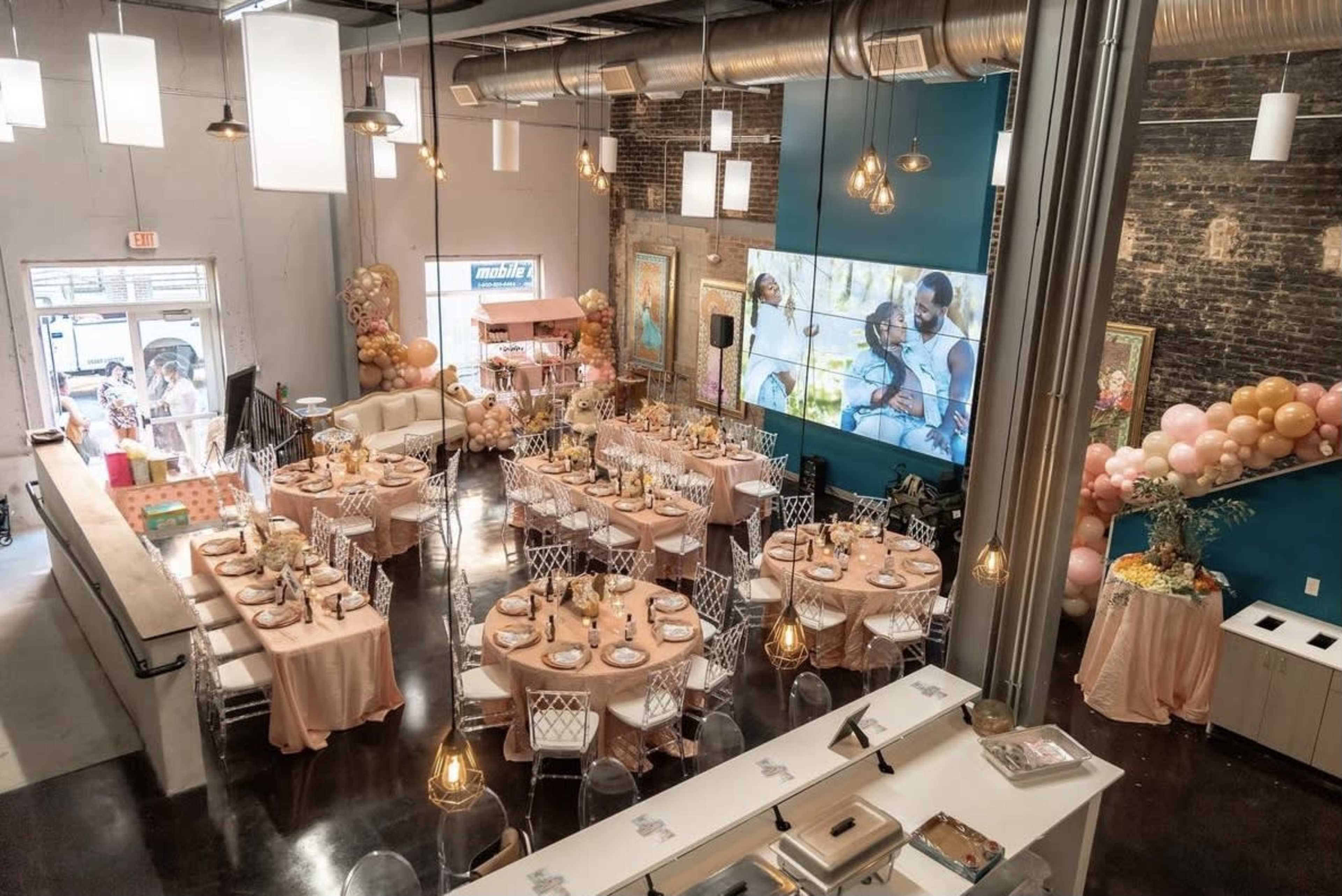 A decorated event space with multiple tables set for a celebration, featuring elegant table settings and a large video screen displaying a scene.
