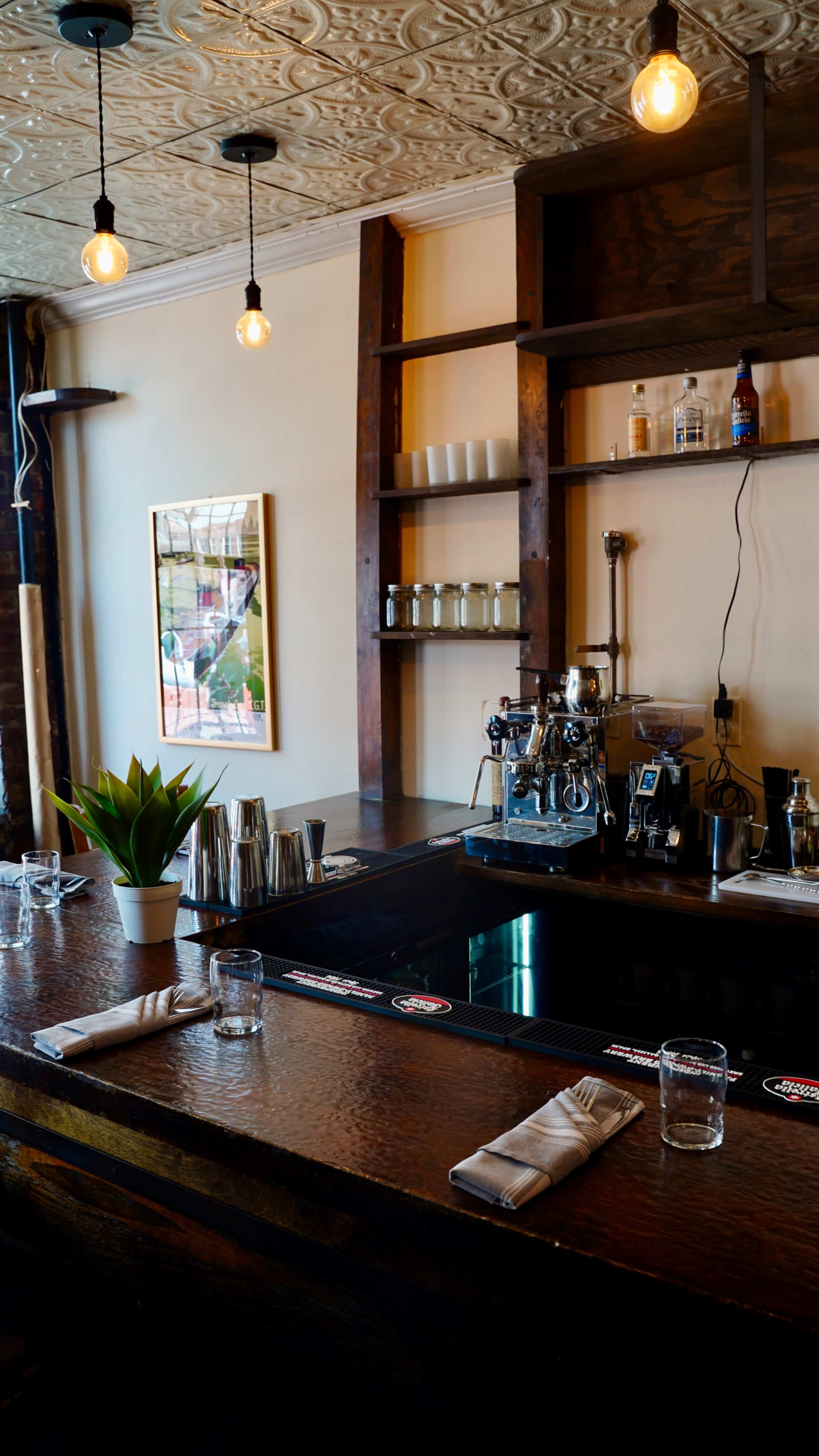 The image shows a bar area with a wooden counter, glassware, and a coffee machine, complemented by shelves displaying drinks and a potted plant.