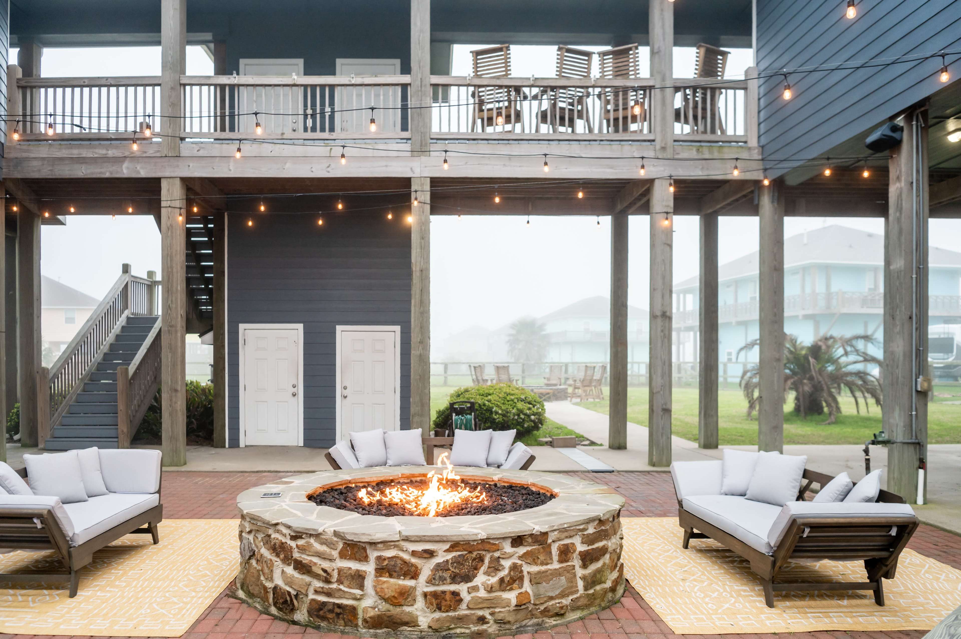A fire pit surrounded by four lounge chairs, situated under a multi-level deck with string lights against a foggy backdrop.