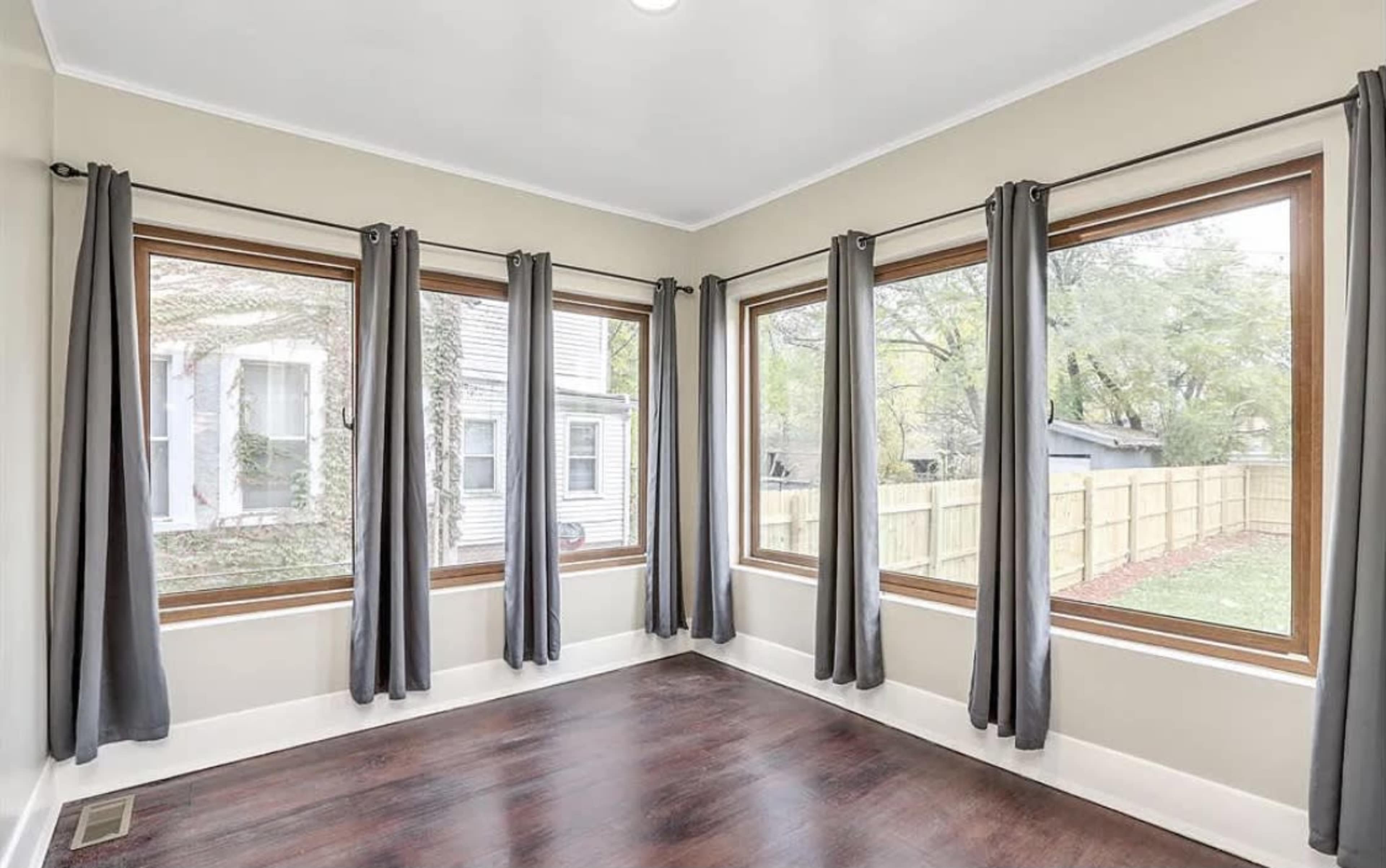 A sunroom with large windows and dark curtains, featuring a wooden floor and light-colored walls.