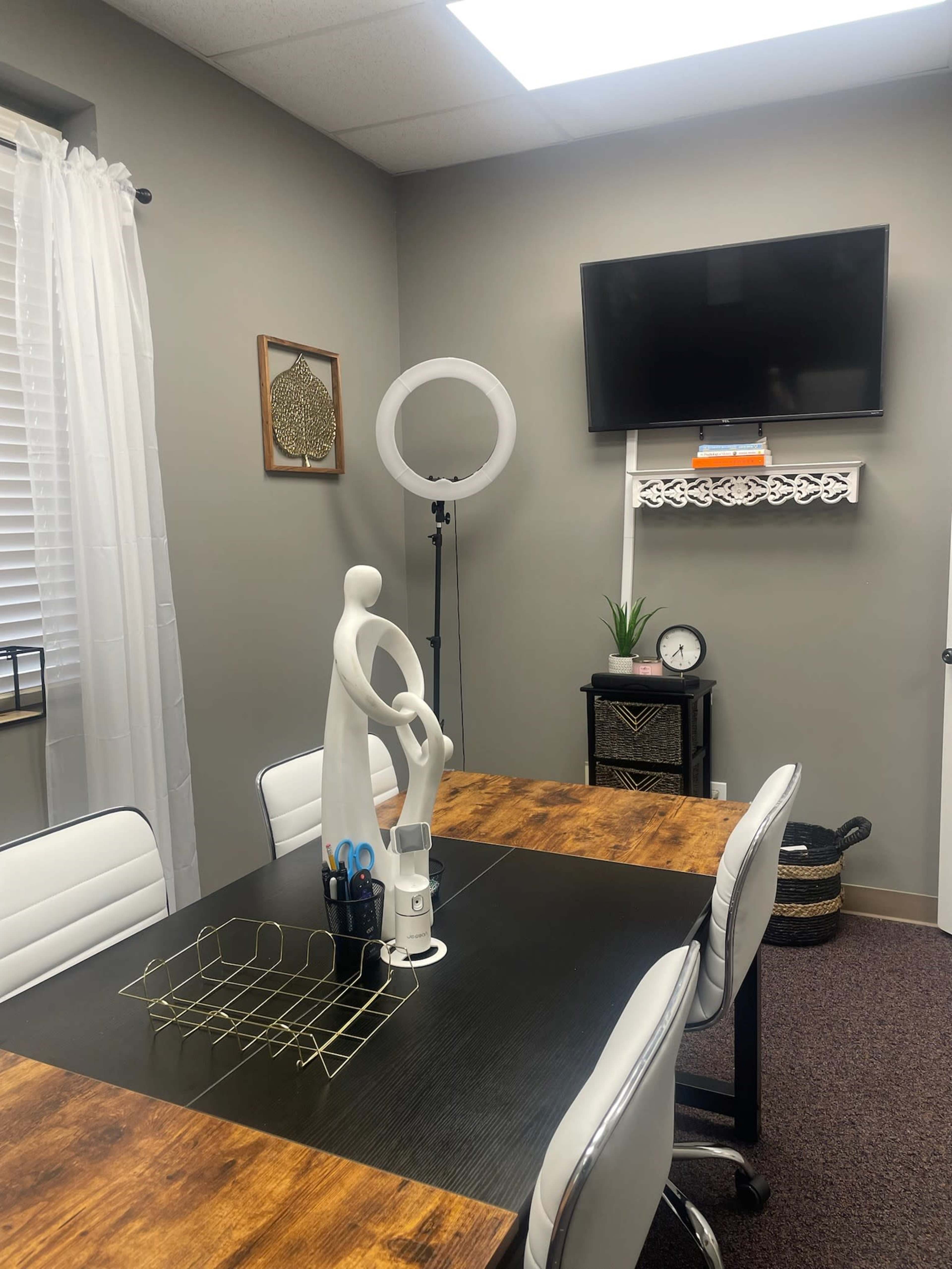A well-lit office space featuring a wooden table surrounded by white chairs, a decorative figure on the table, and a wall-mounted TV above a shelf.