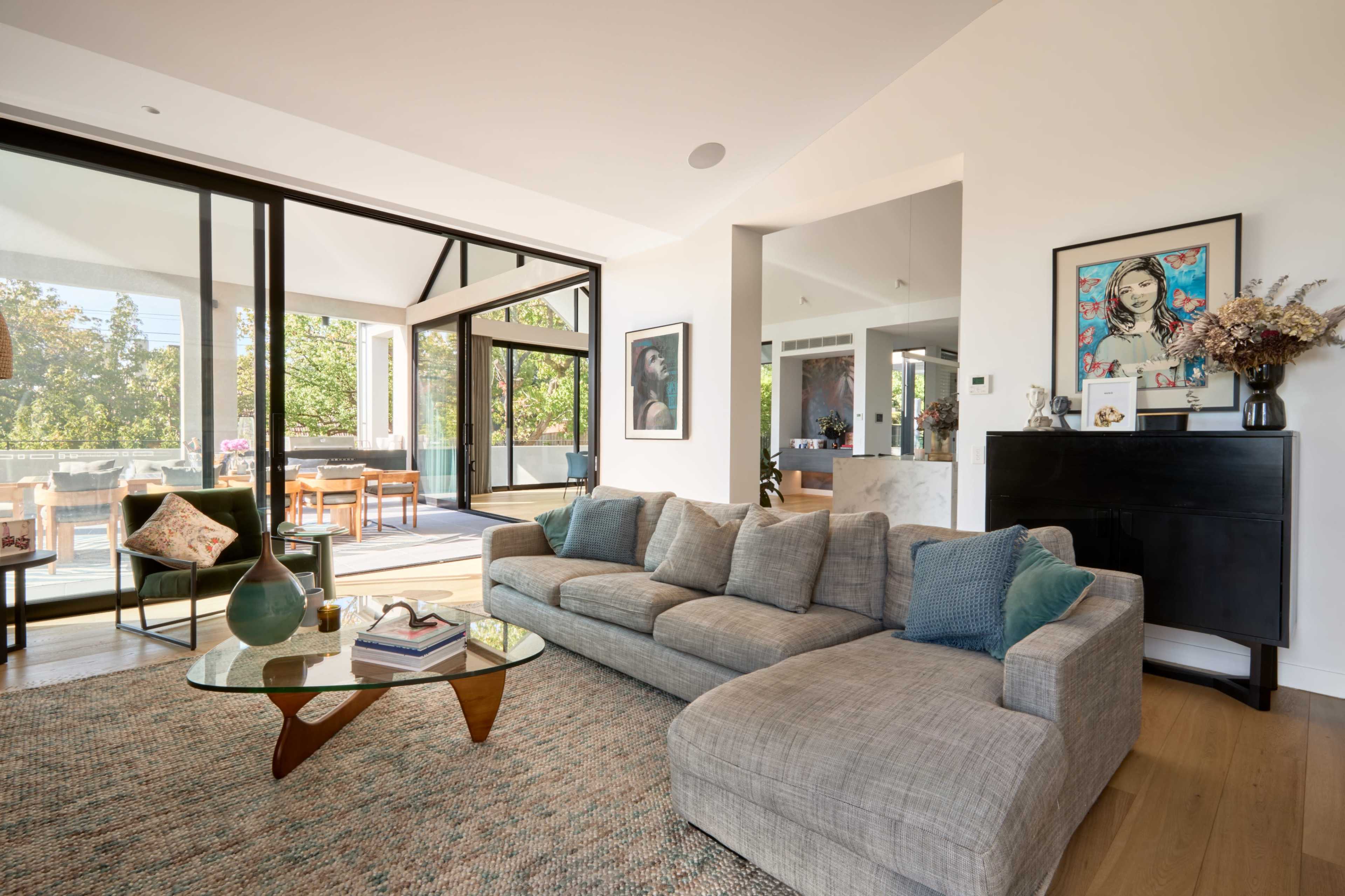 The image shows a modern living room with a gray sectional sofa, a glass coffee table, and large windows providing a view of greenery outside.