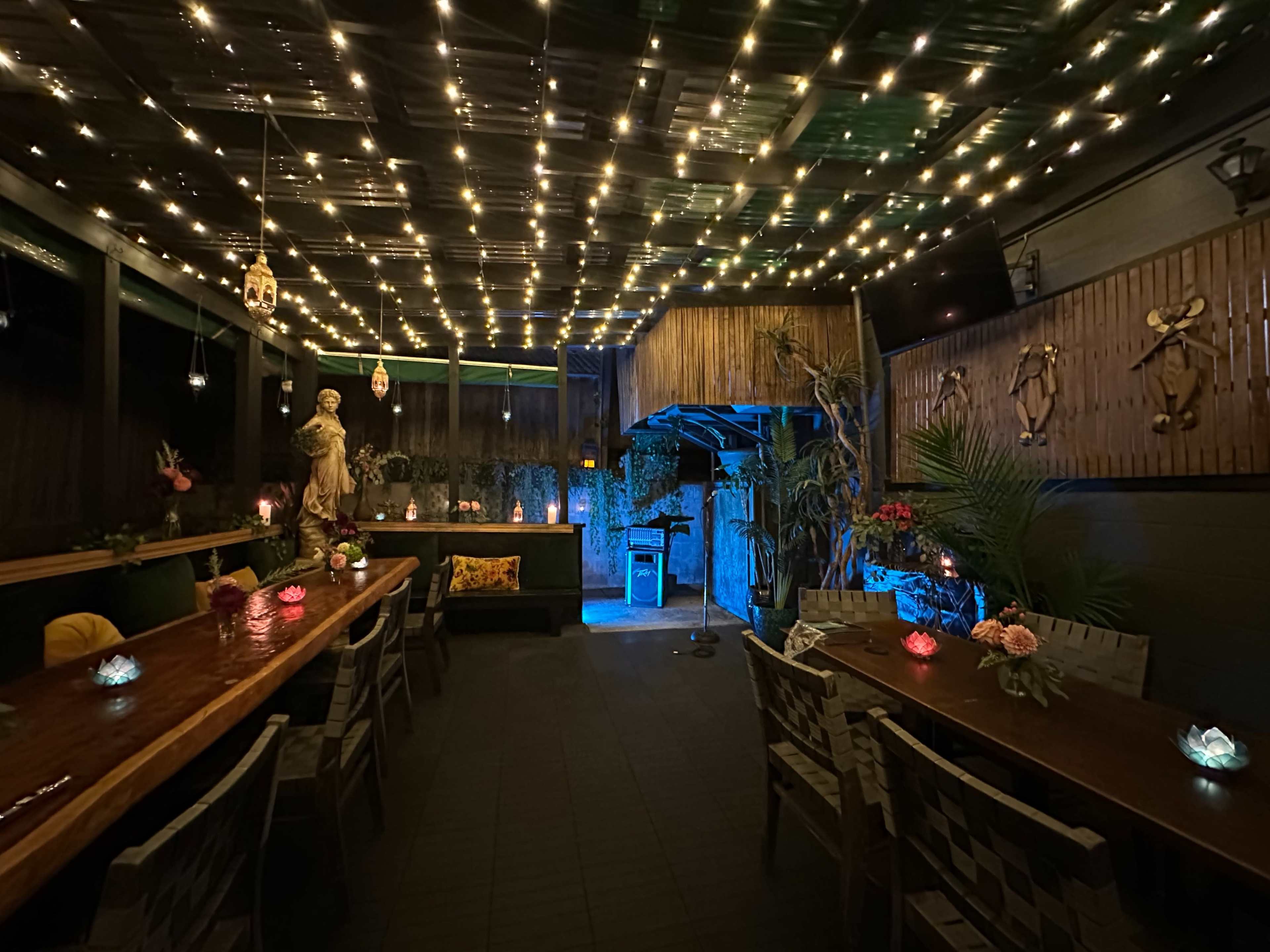 The image shows an illuminated outdoor dining area with wooden tables adorned with colorful decorative lights and plants, under a ceiling of string lights.