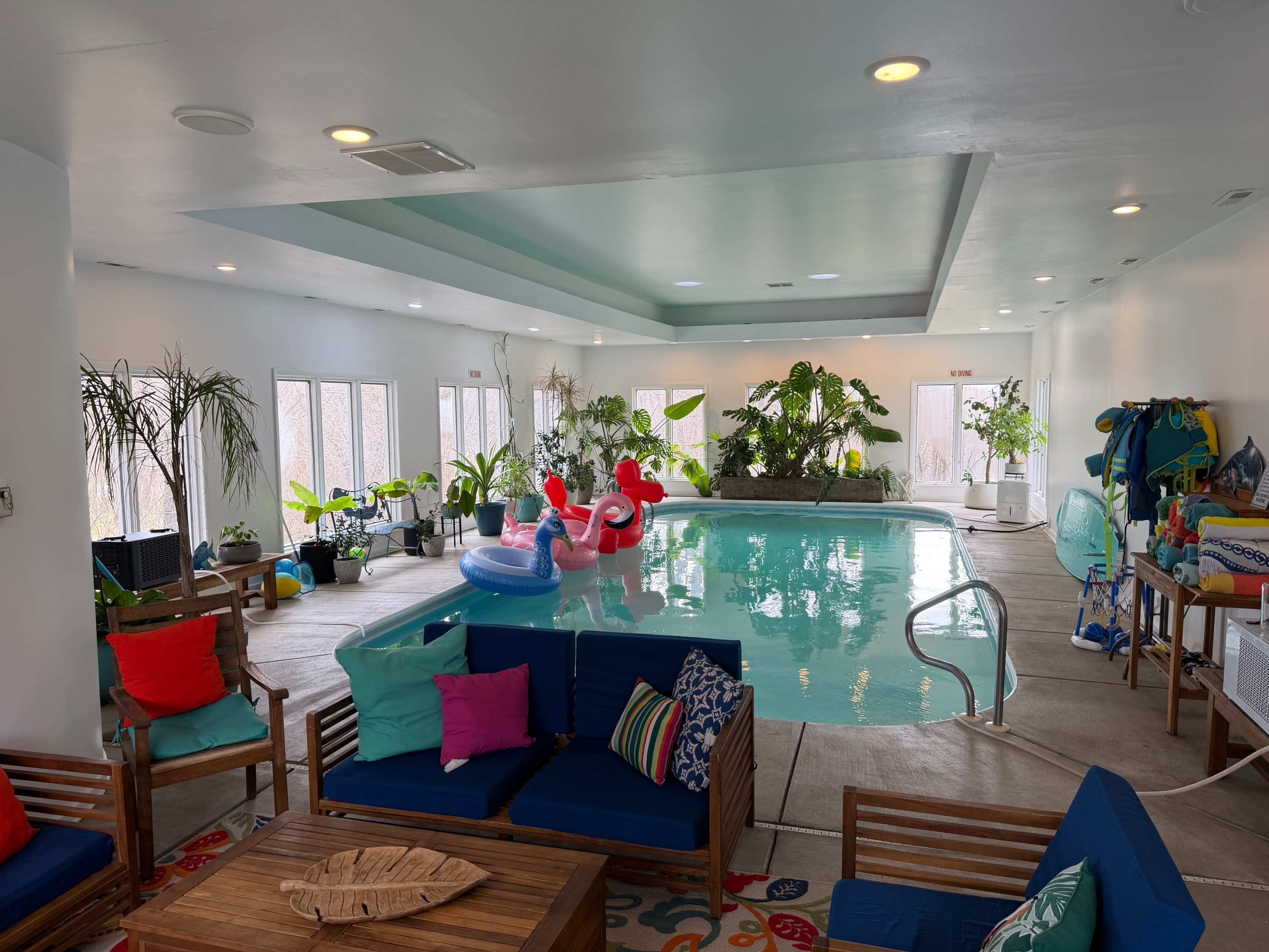 The image shows an indoor swimming pool surrounded by plants and lounge seating, with colorful inflatable toys floating in the water.
