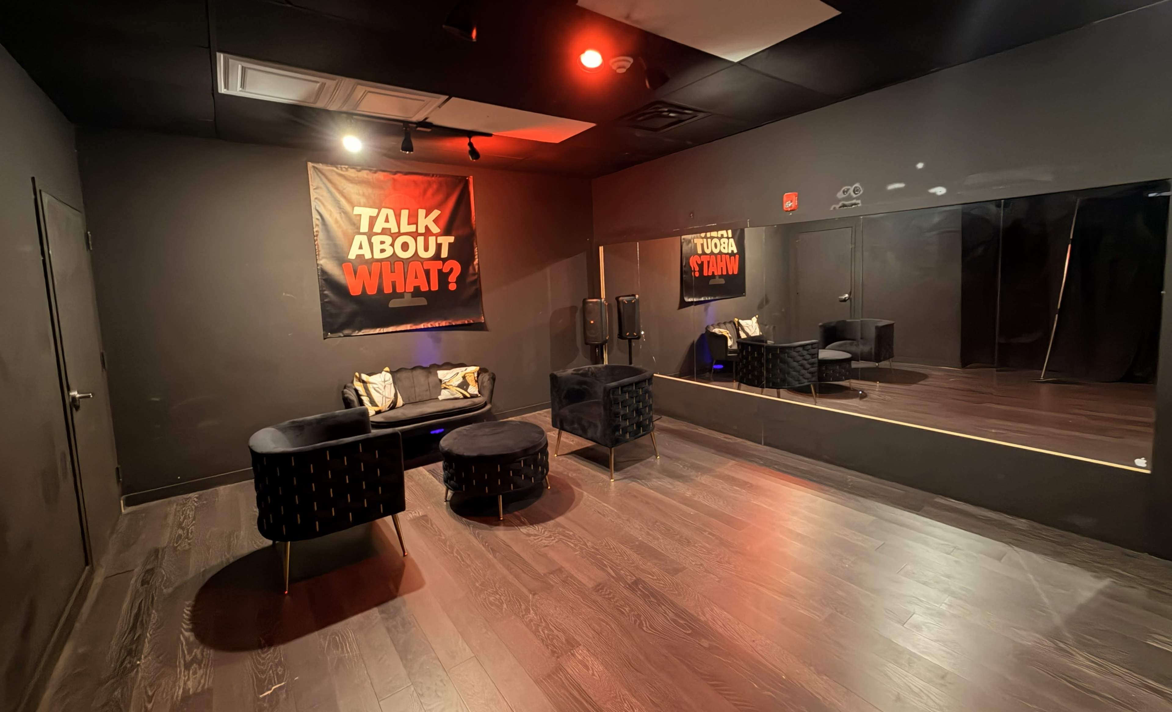 The image shows a small, dark room with black walls, featuring a large poster that reads "TALK ABOUT WHAT?" and two round, black chairs on a hardwood floor.