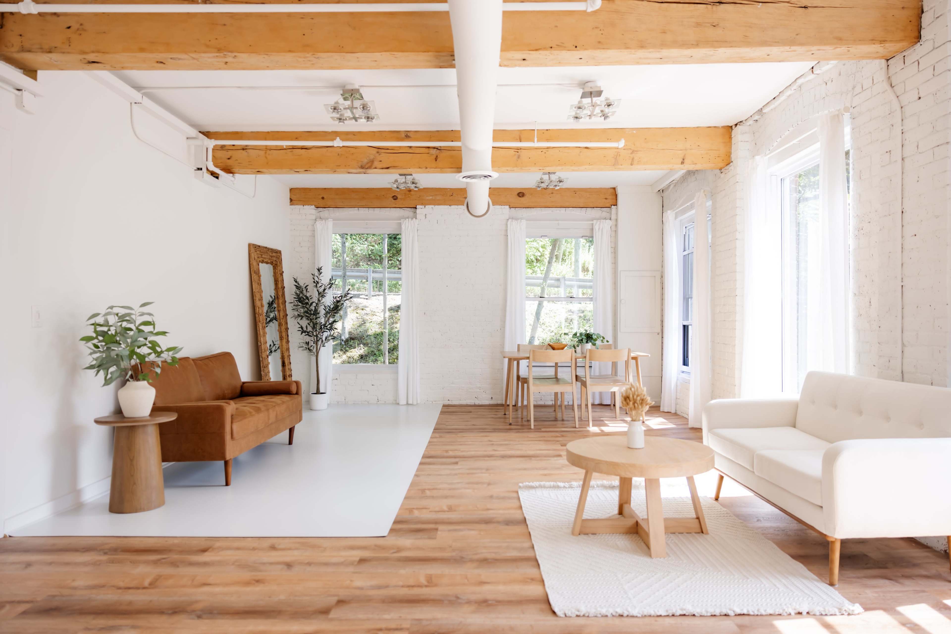 The image shows a bright, modern interior with wooden beams, a sofa, a coffee table, and dining area, all set against white brick walls.