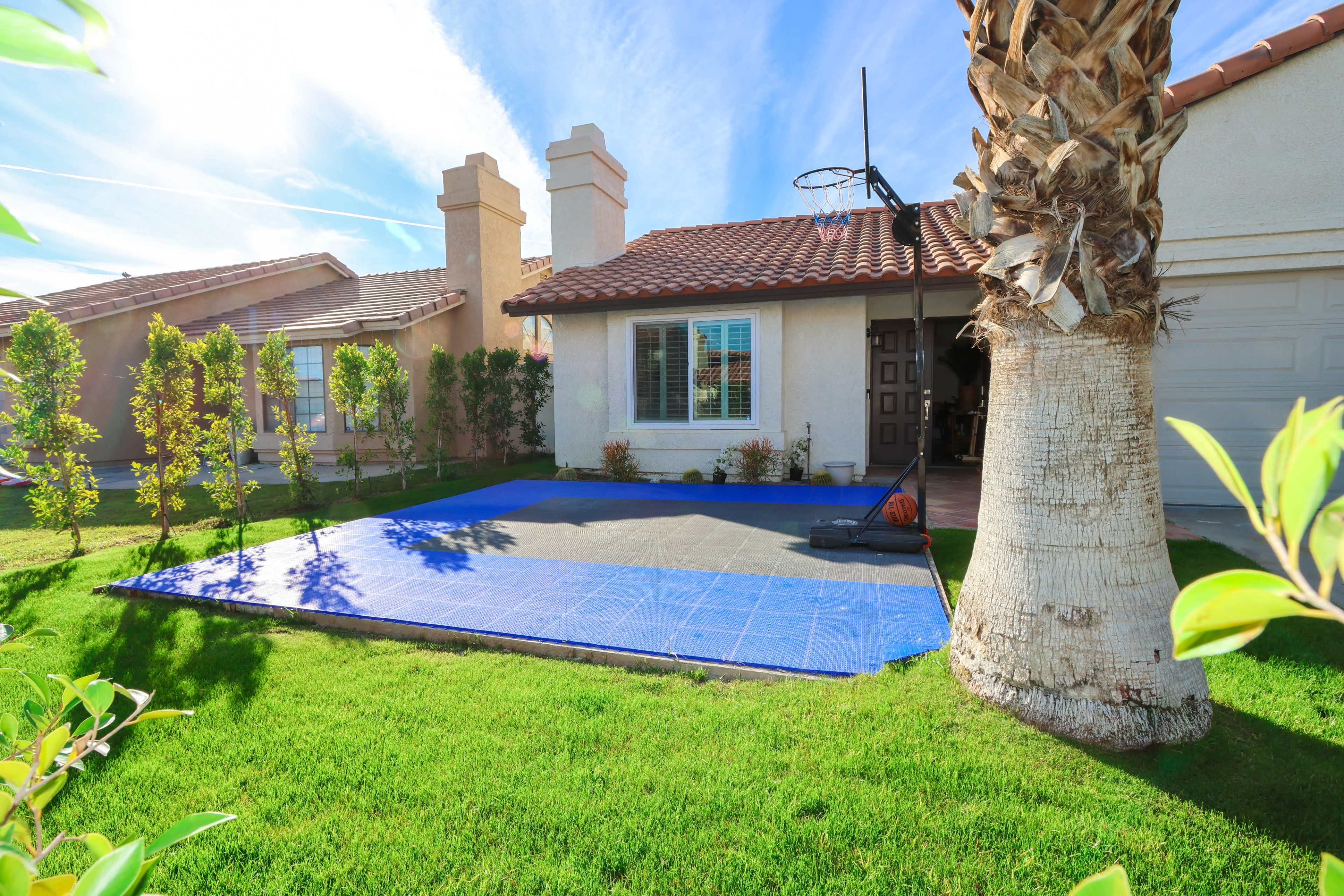 The image shows a residential home with a basketball hoop and a blue court area in the front yard, surrounded by green grass and landscaping.