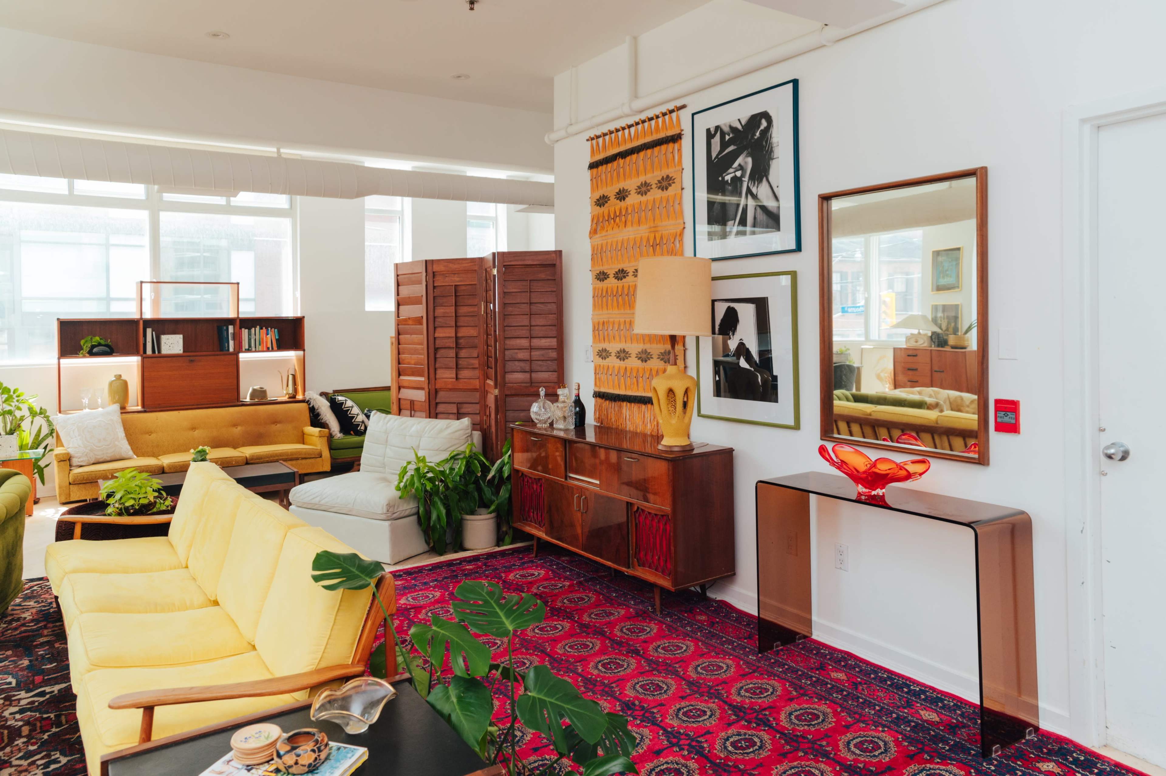A bright living room with a mix of mid-century modern furniture, including a yellow sofa, a wooden sideboard, and various decorative plants.