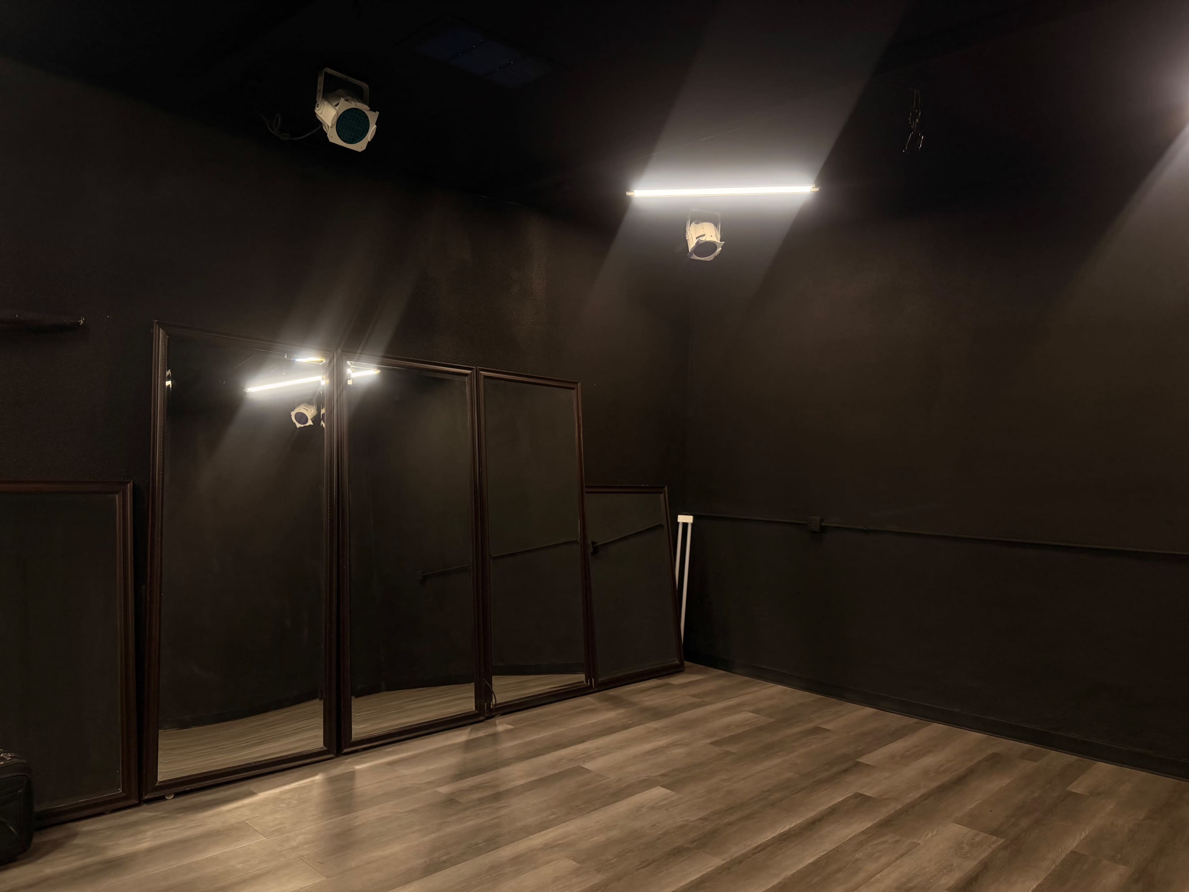 The image shows a dimly lit rehearsal space with a dark wall, wooden floor, and a set of three mirrors on one side.