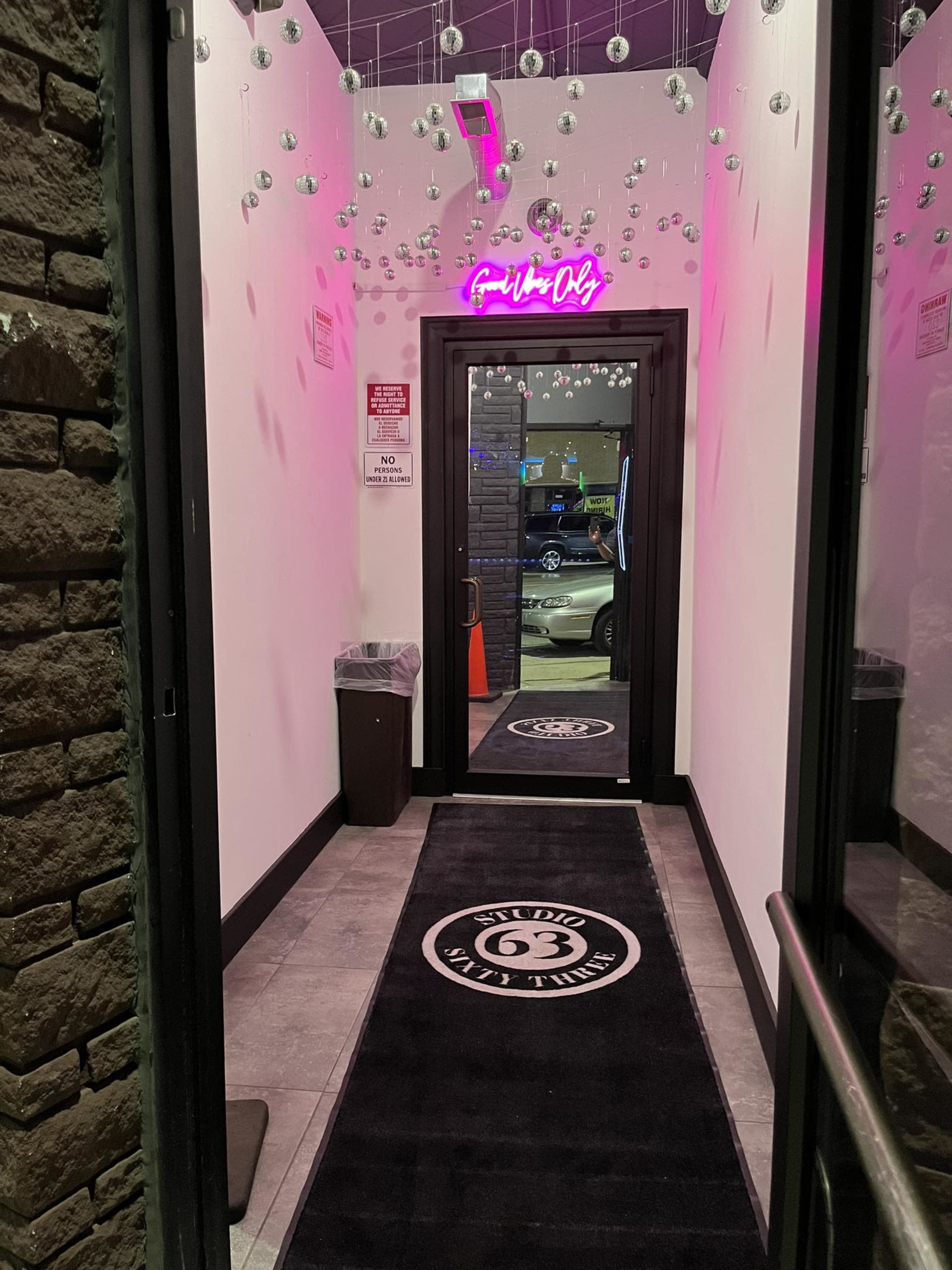 A narrow hallway with a black carpet featuring a logo, illuminated by pink neon lights and adorned with hanging decorative elements.