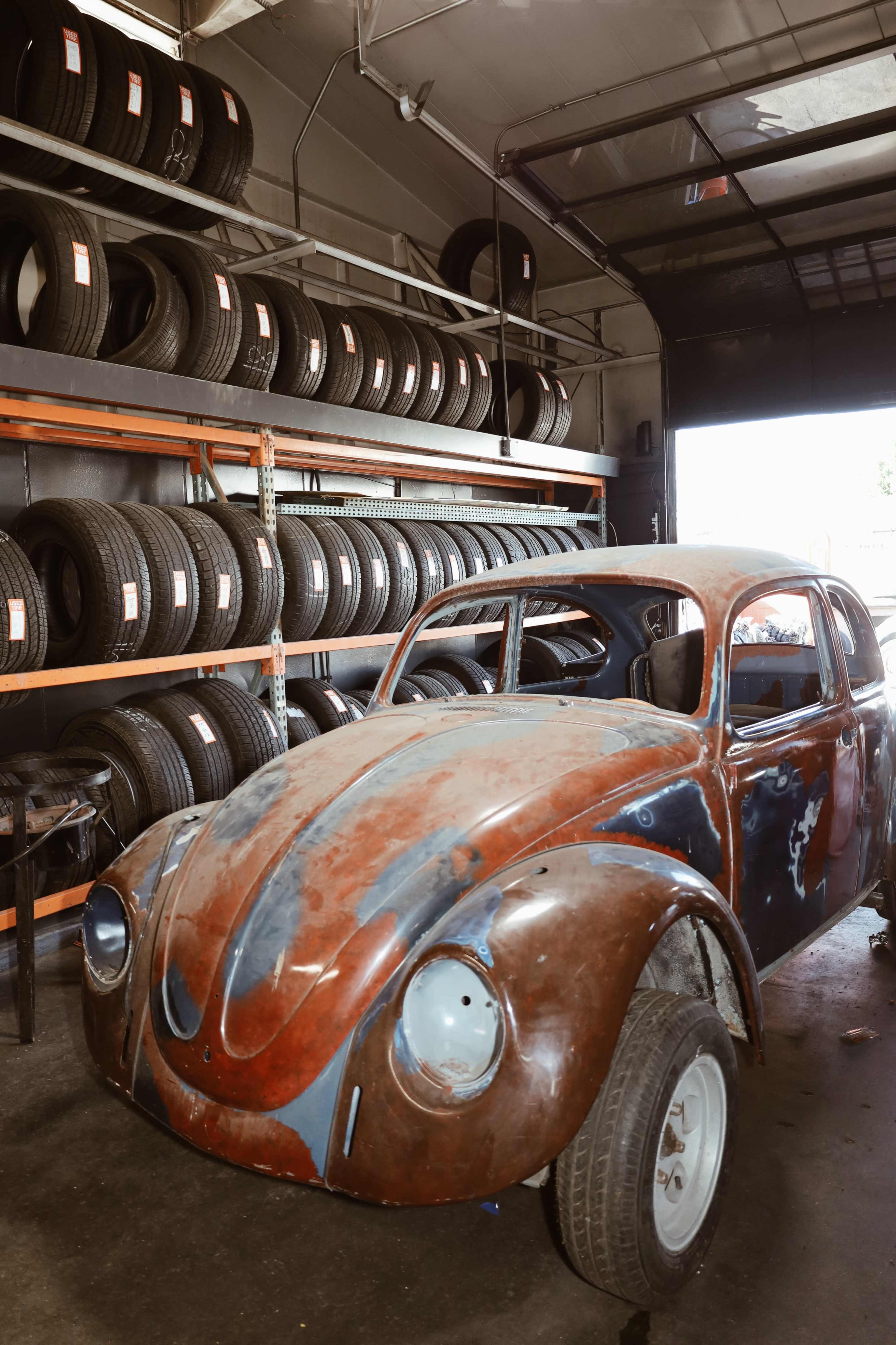 A vintage car with a weathered body is parked in a garage next to a wall stacked with tires.