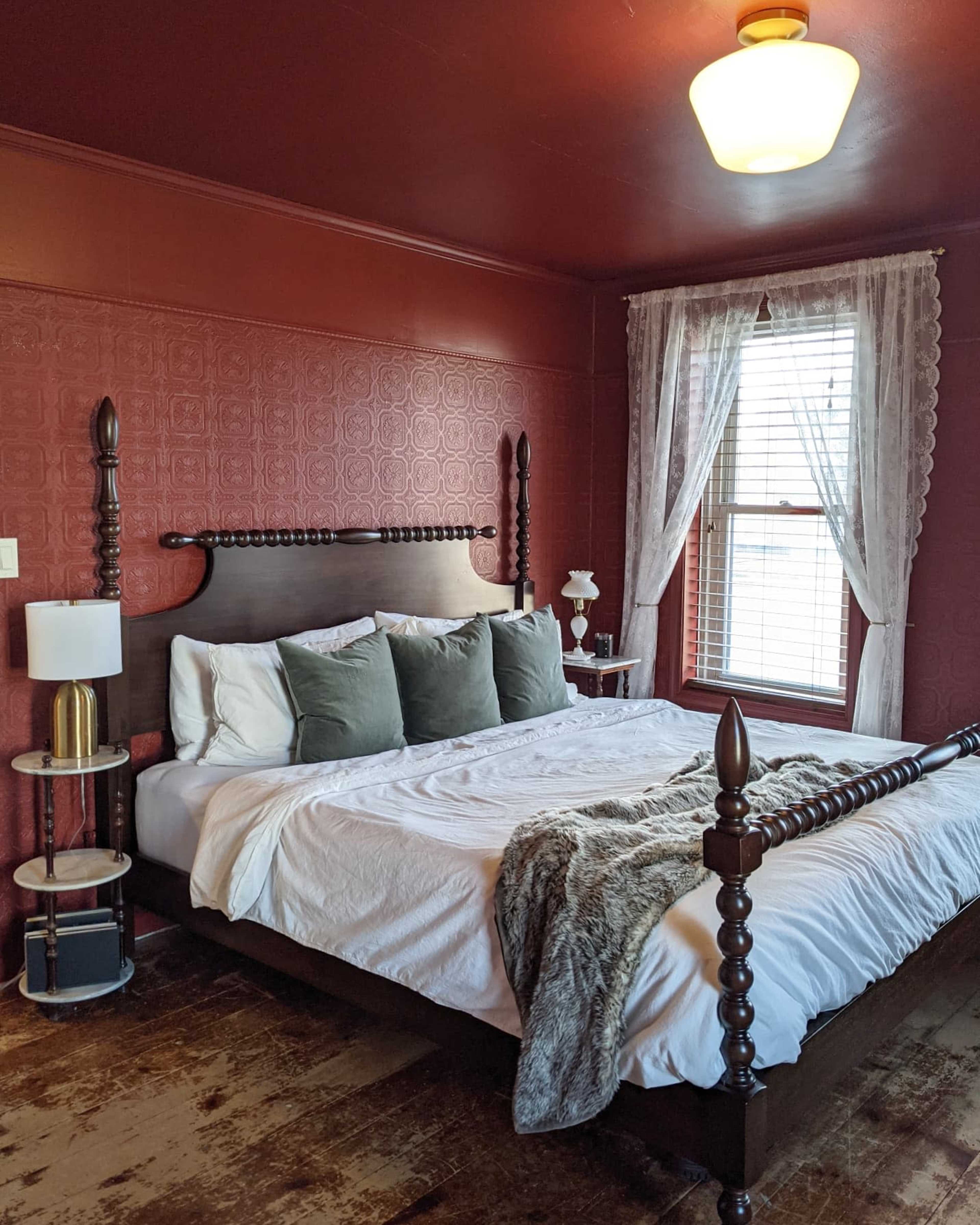 A cozy bedroom with a dark red feature wall, a wooden bed with ornate posts, and light-colored bedding, paired with a bedside table and a window adorned with white curtains.