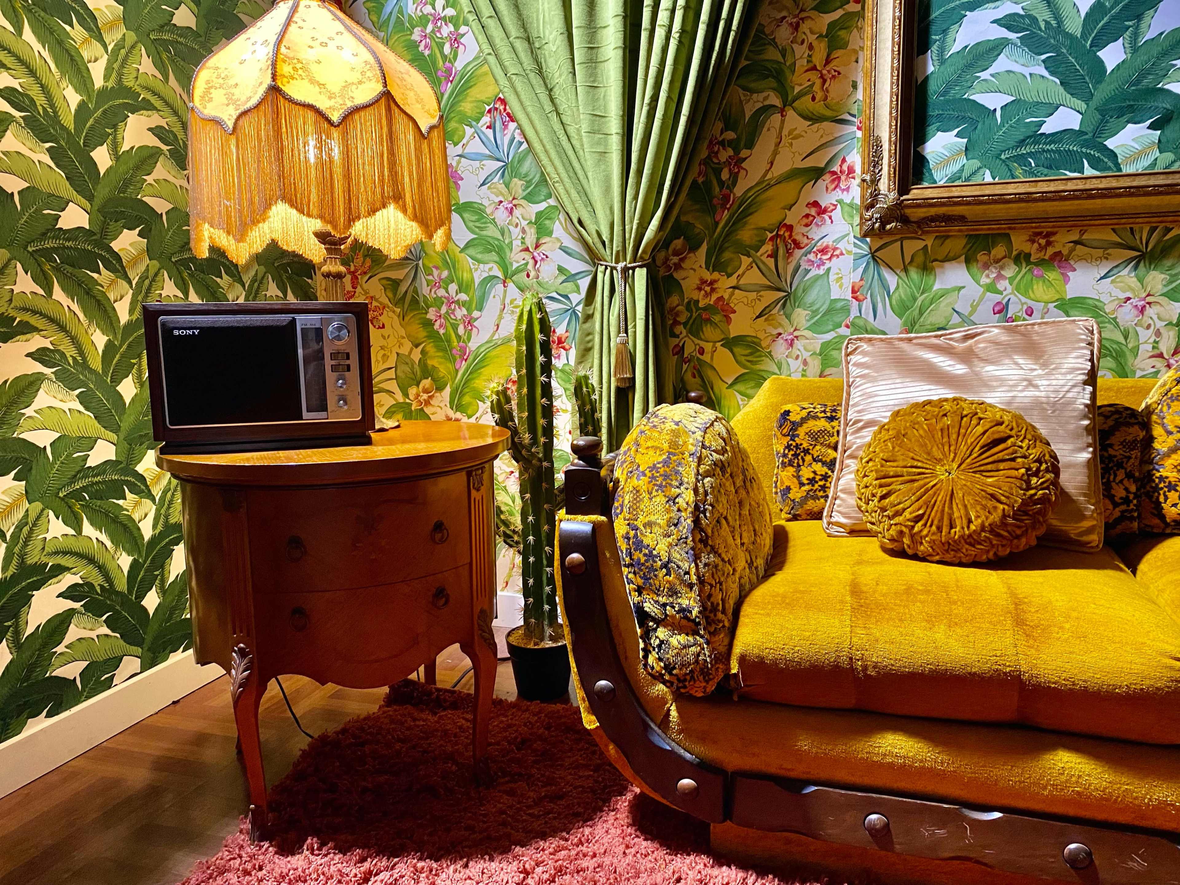 A vintage living room setting with a yellow sofa, a round wooden table holding a microwave, and a large ornate lamp, all surrounded by tropical-patterned wallpaper and curtains.