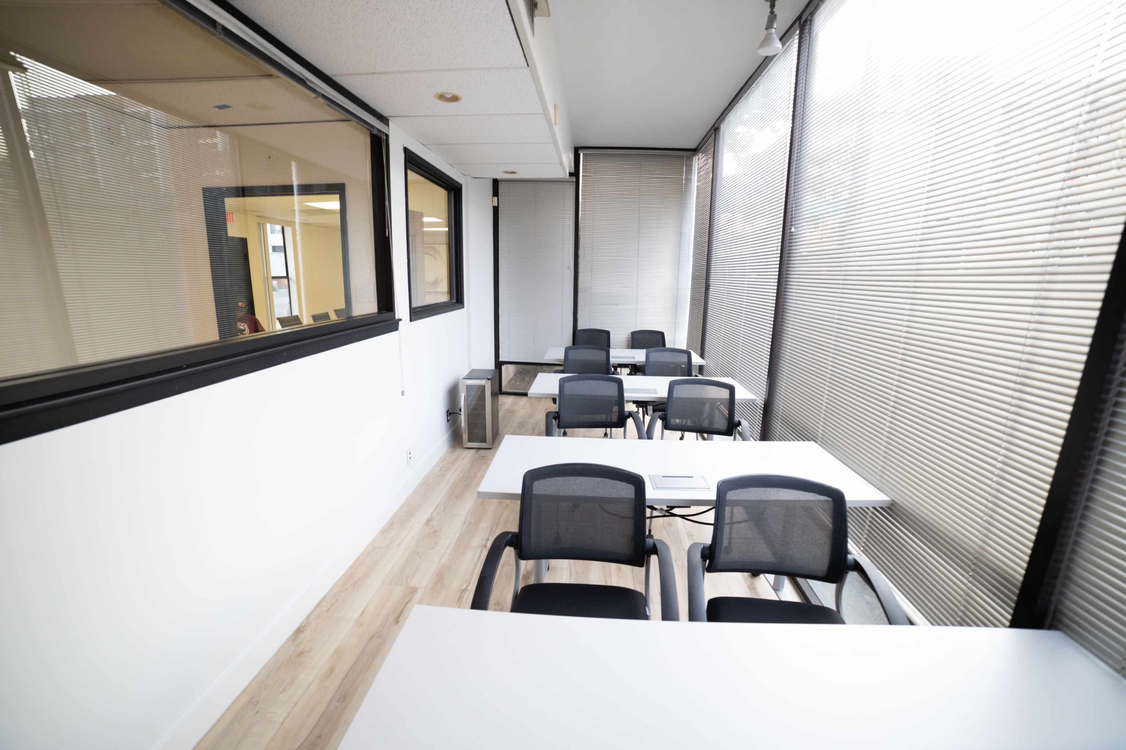 A bright conference room features several tables and chairs arranged along a wall of large windows with blinds.