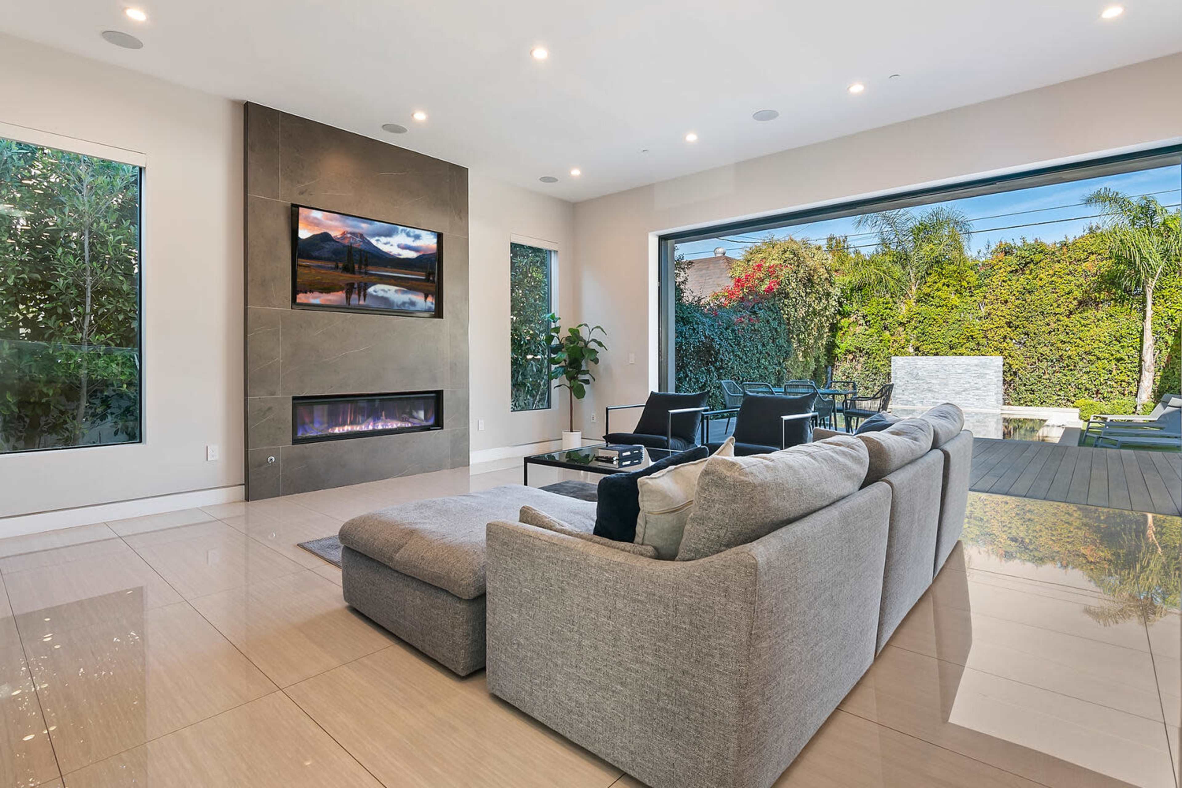 A modern living room with a large sectional sofa facing a wall-mounted television, large windows, and a view of a landscaped outdoor area.