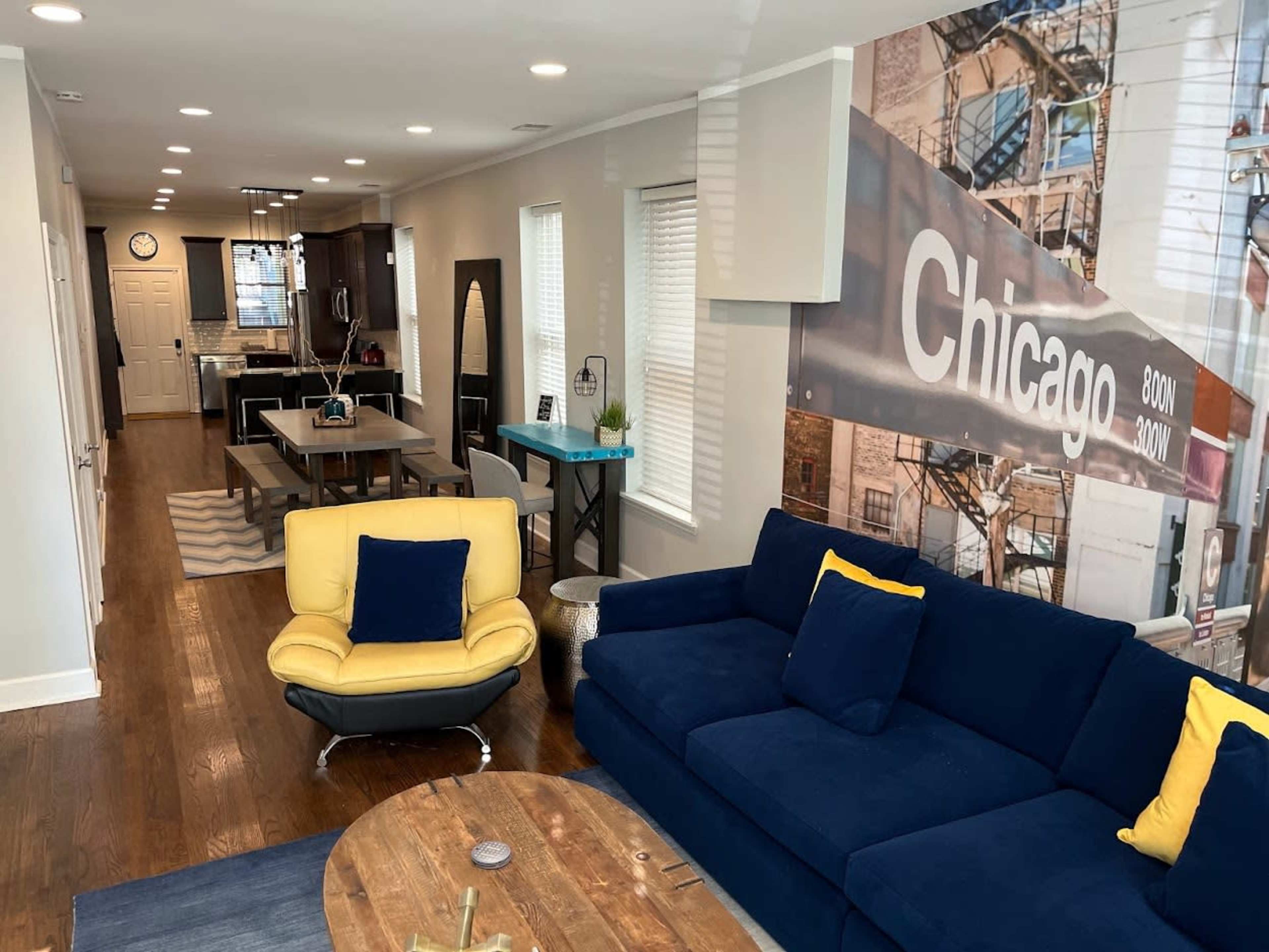 A modern living room with a blue sofa and a yellow chair, leading to a dining area and kitchen in the background, with a large wall mural of Chicago.