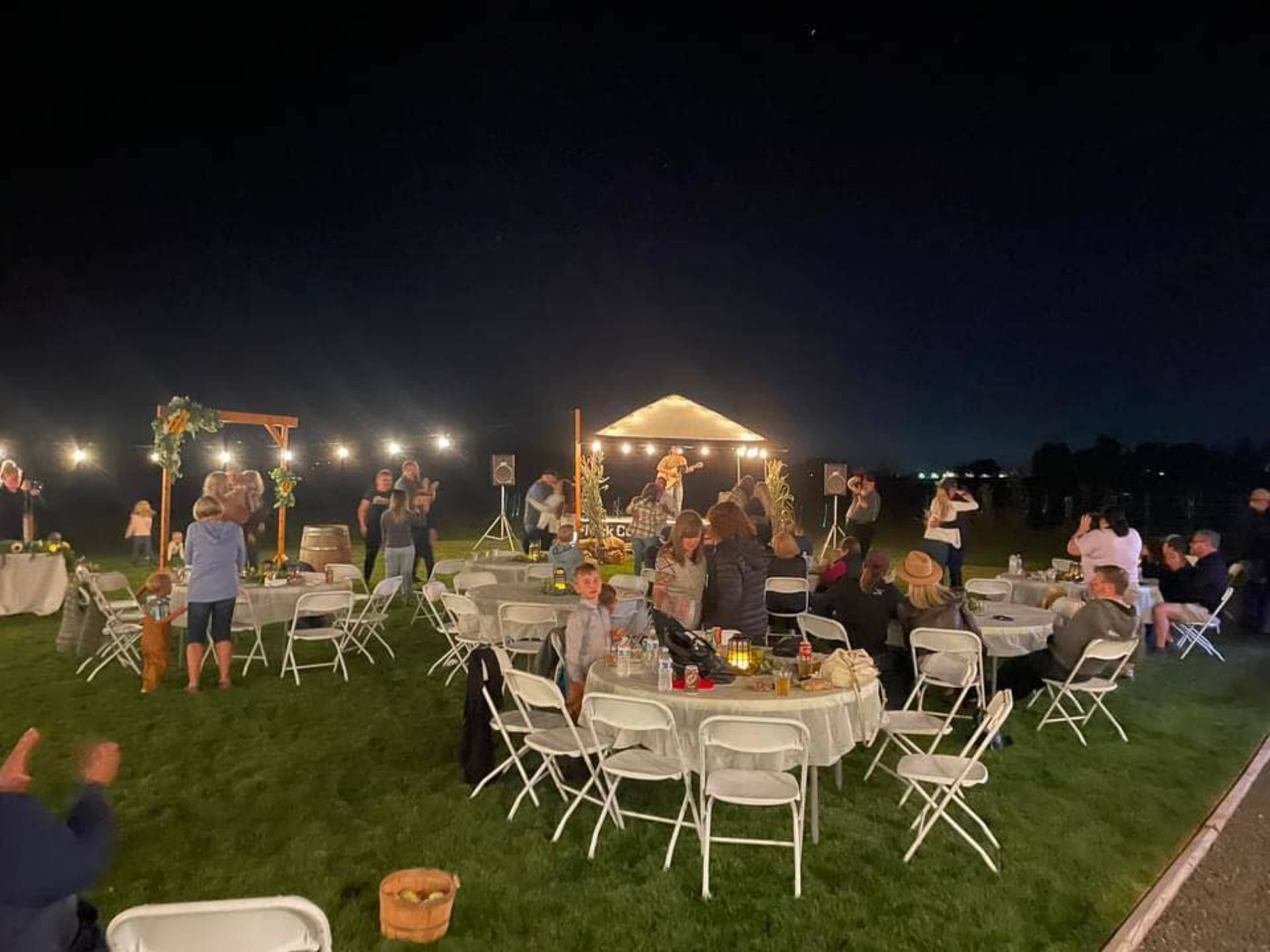 A large outdoor event features numerous white tables and chairs, with guests socializing under string lights at night.