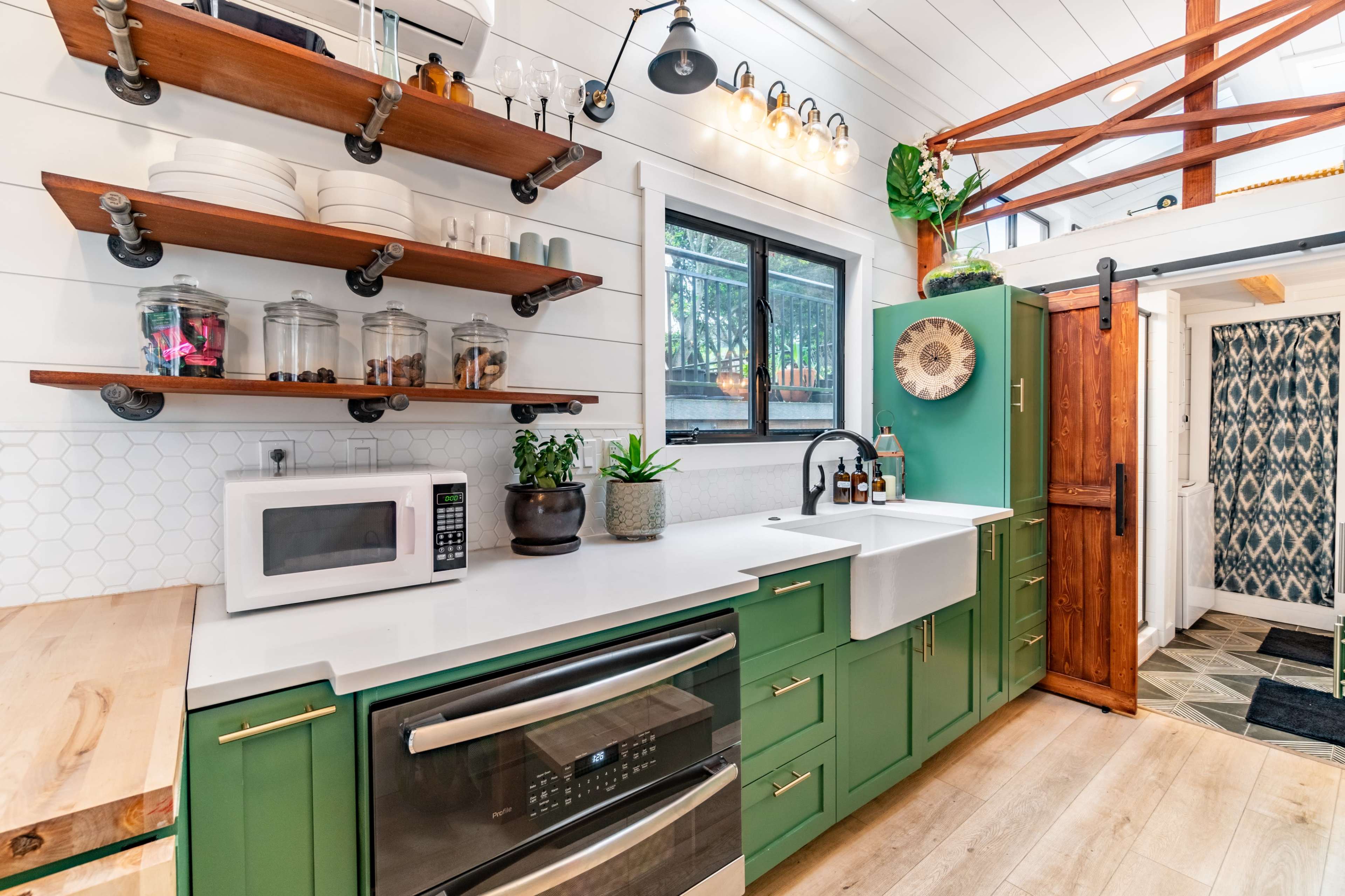 A compact kitchen featuring green cabinetry, open wood shelves with dishware, a farmhouse sink, and a window overlooking a garden.