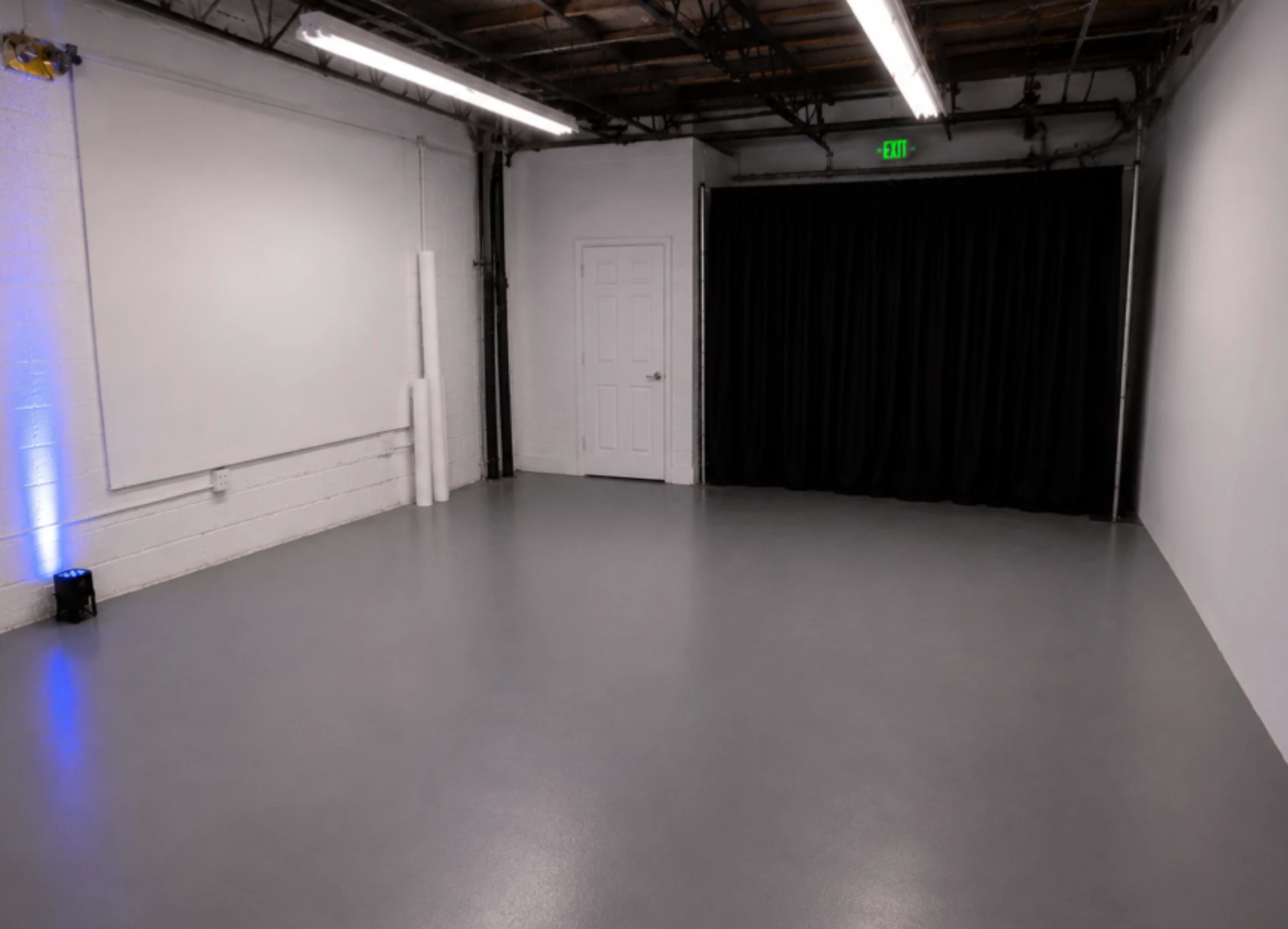 The image shows a large, empty studio space with gray flooring, a white wall, and a black curtain covering a doorway.