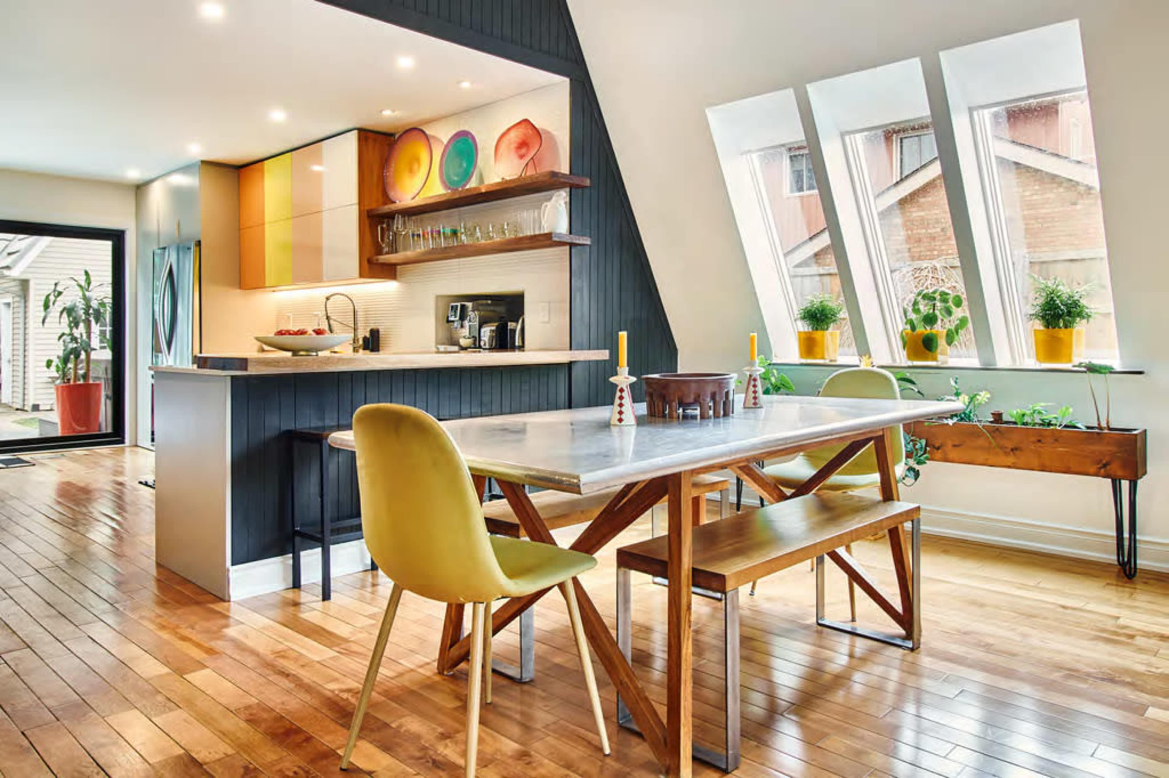 A modern kitchen-dining area features a marble table surrounded by wooden benches and yellow chairs, with large angled windows and colorful dishware displayed on the wall.