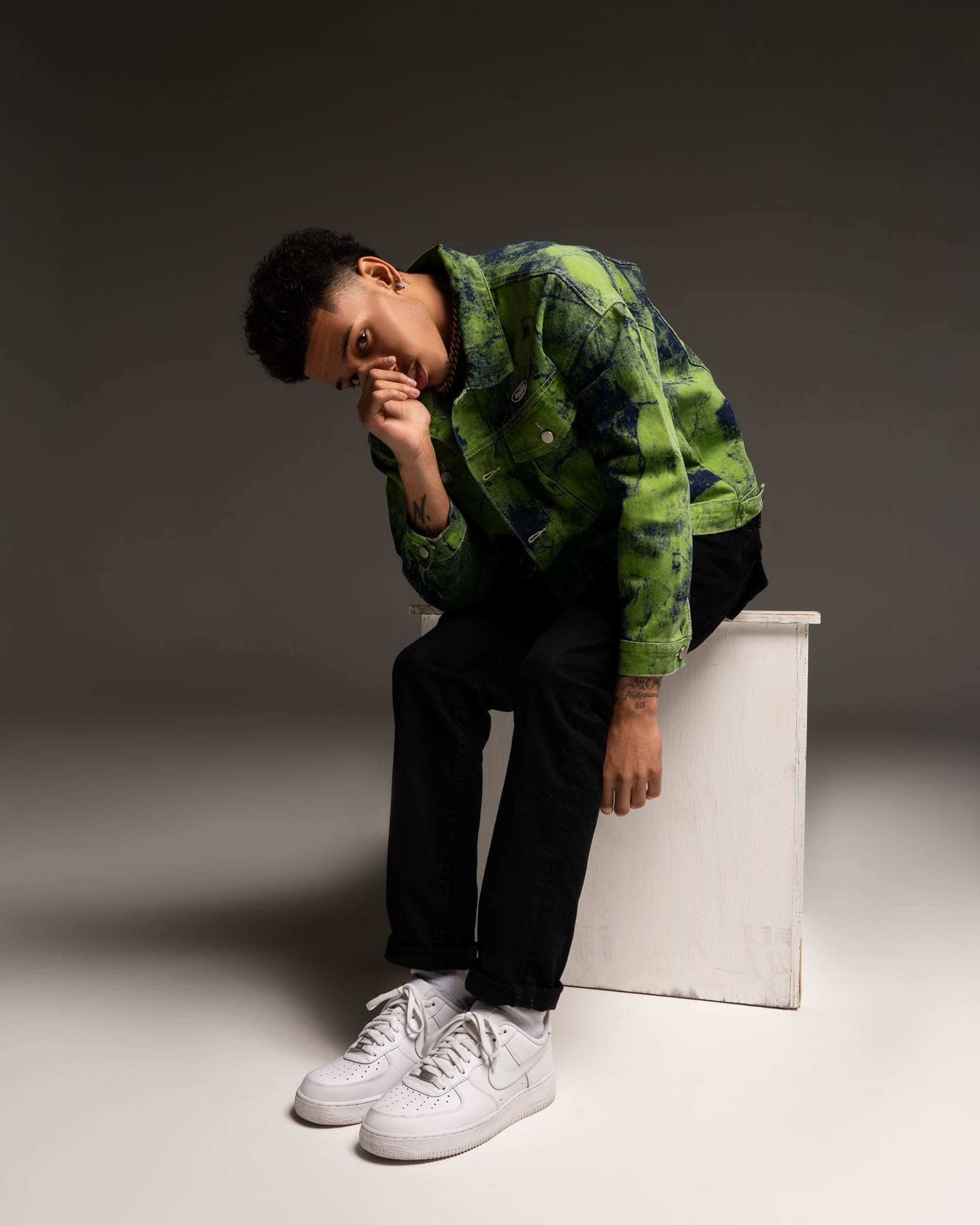 A person sits on a white stool, wearing a green and black patterned jacket, black pants, and white sneakers, with their hand covering their mouth.