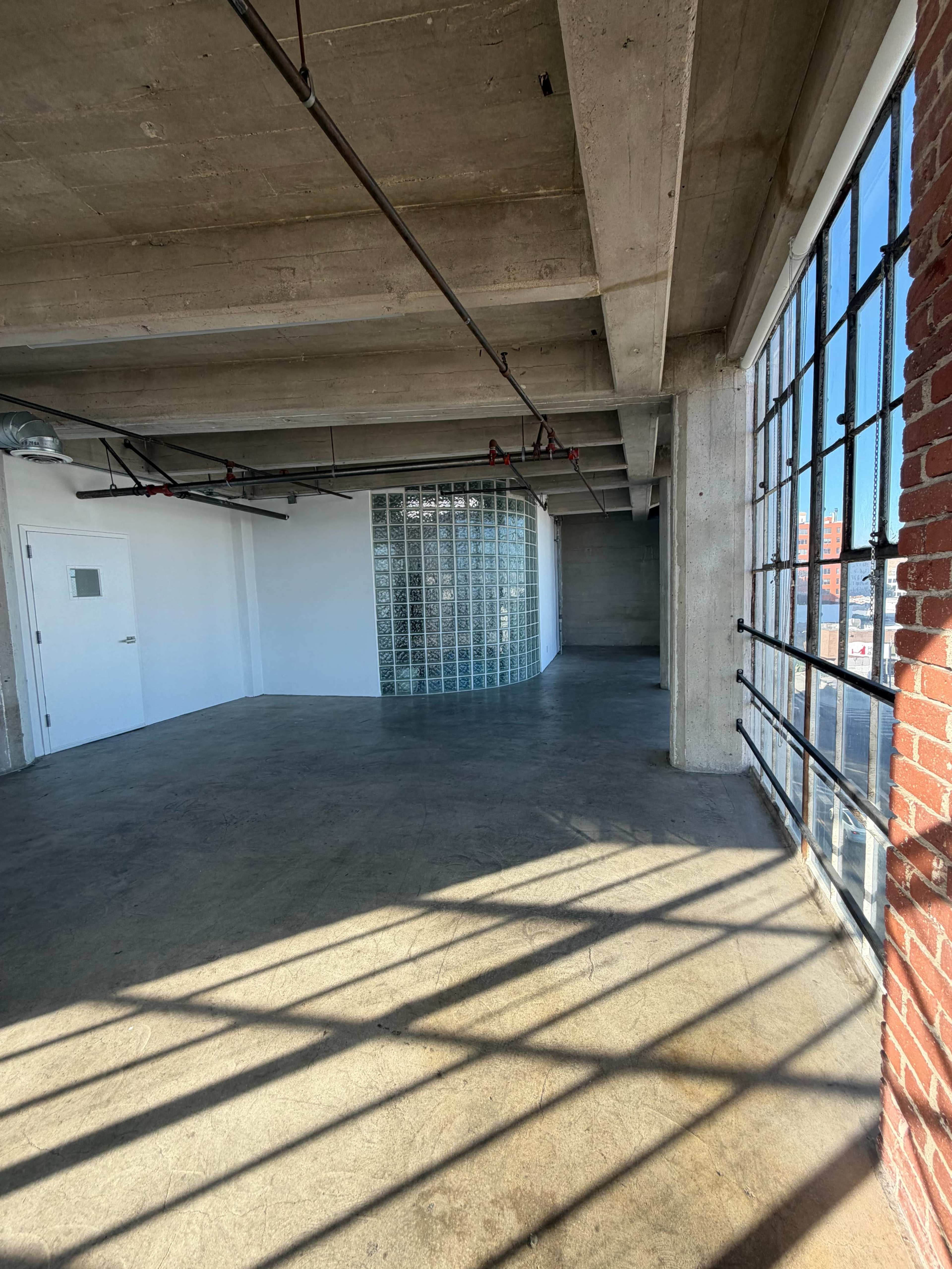 The image shows a spacious, industrial-style room with concrete walls and floors, featuring large windows that allow natural light to enter and a glass block partition in the background.