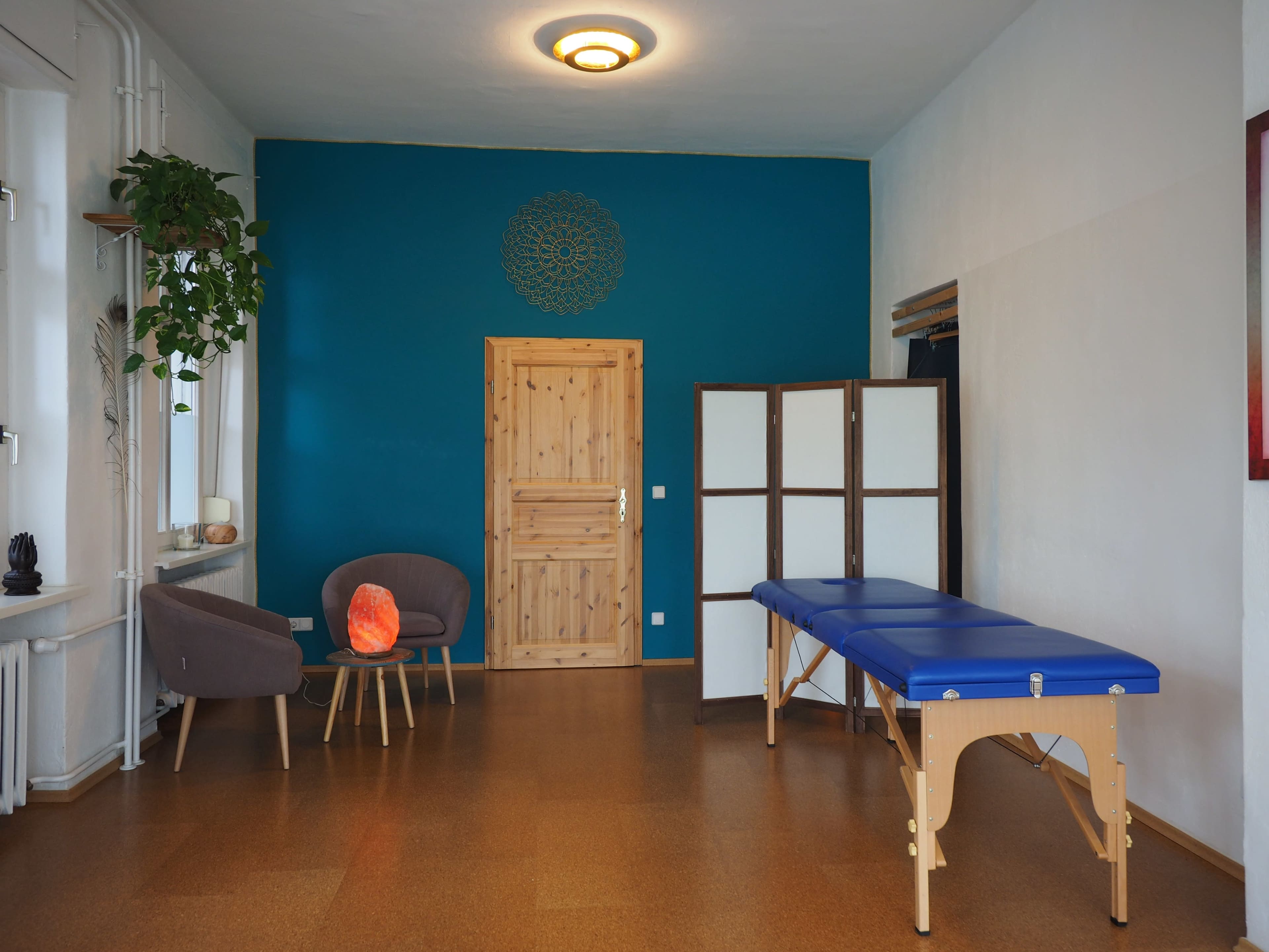 The image shows a spacious room with a blue accent wall, a wooden door, a massage table, a chair, and a plant.