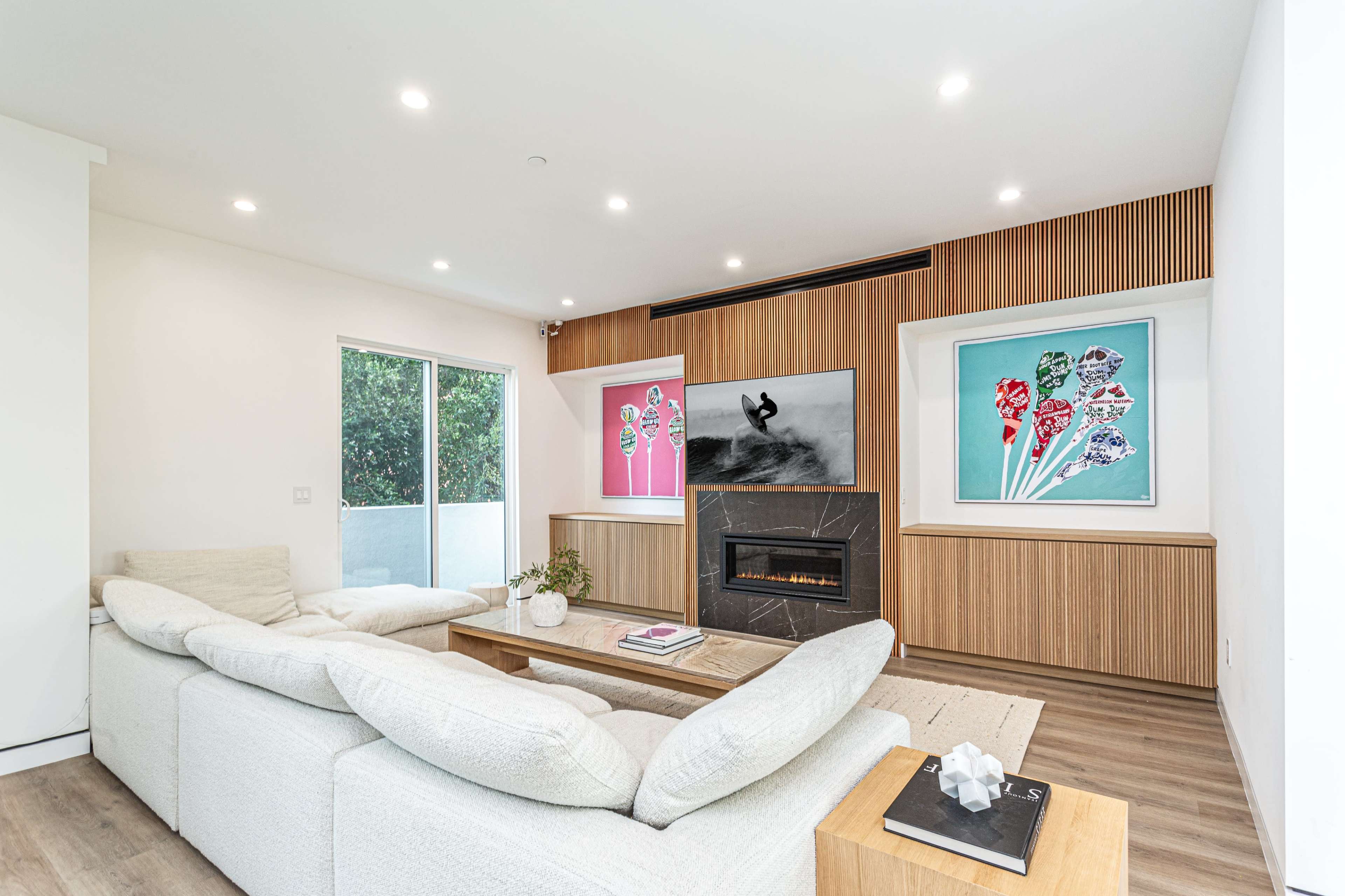 A modern living room features a light-colored sectional sofa, a wooden wall with framed artworks, and a central fireplace.