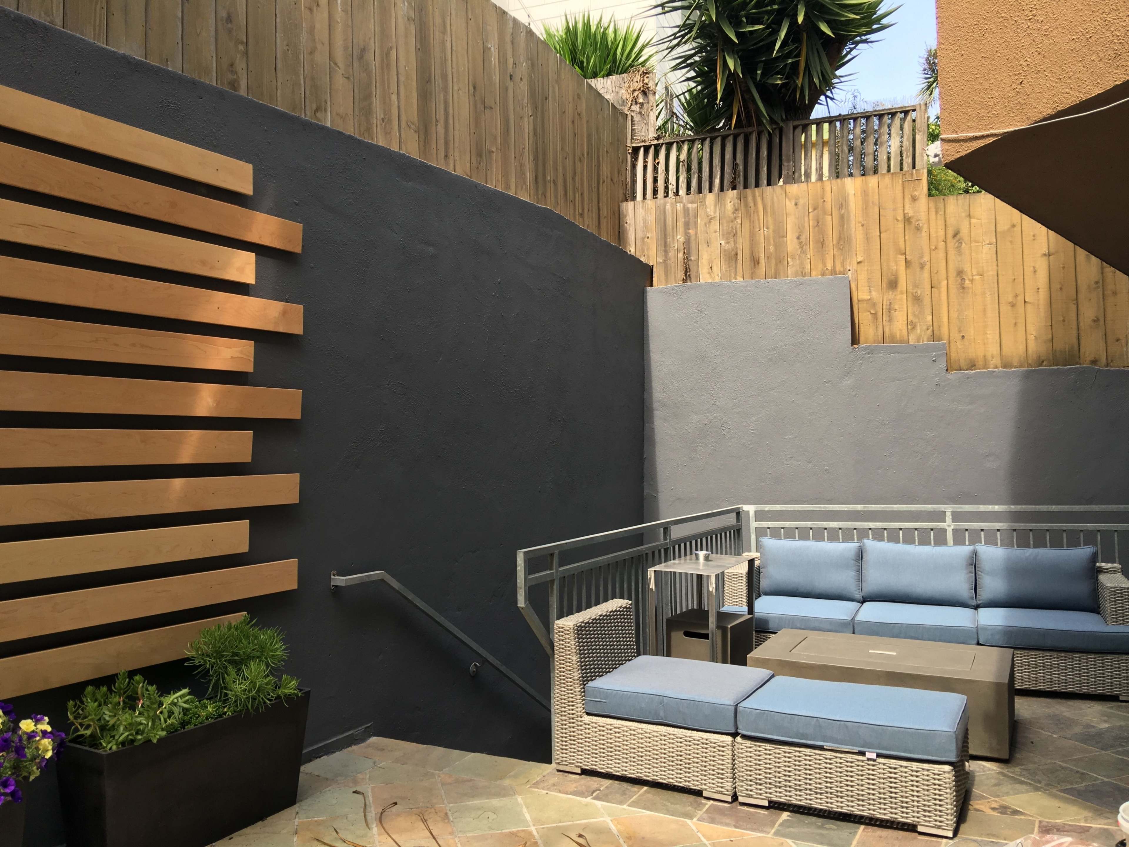 The image shows a modern outdoor seating area with a gray wall, wooden slats, and blue cushions on wicker furniture.