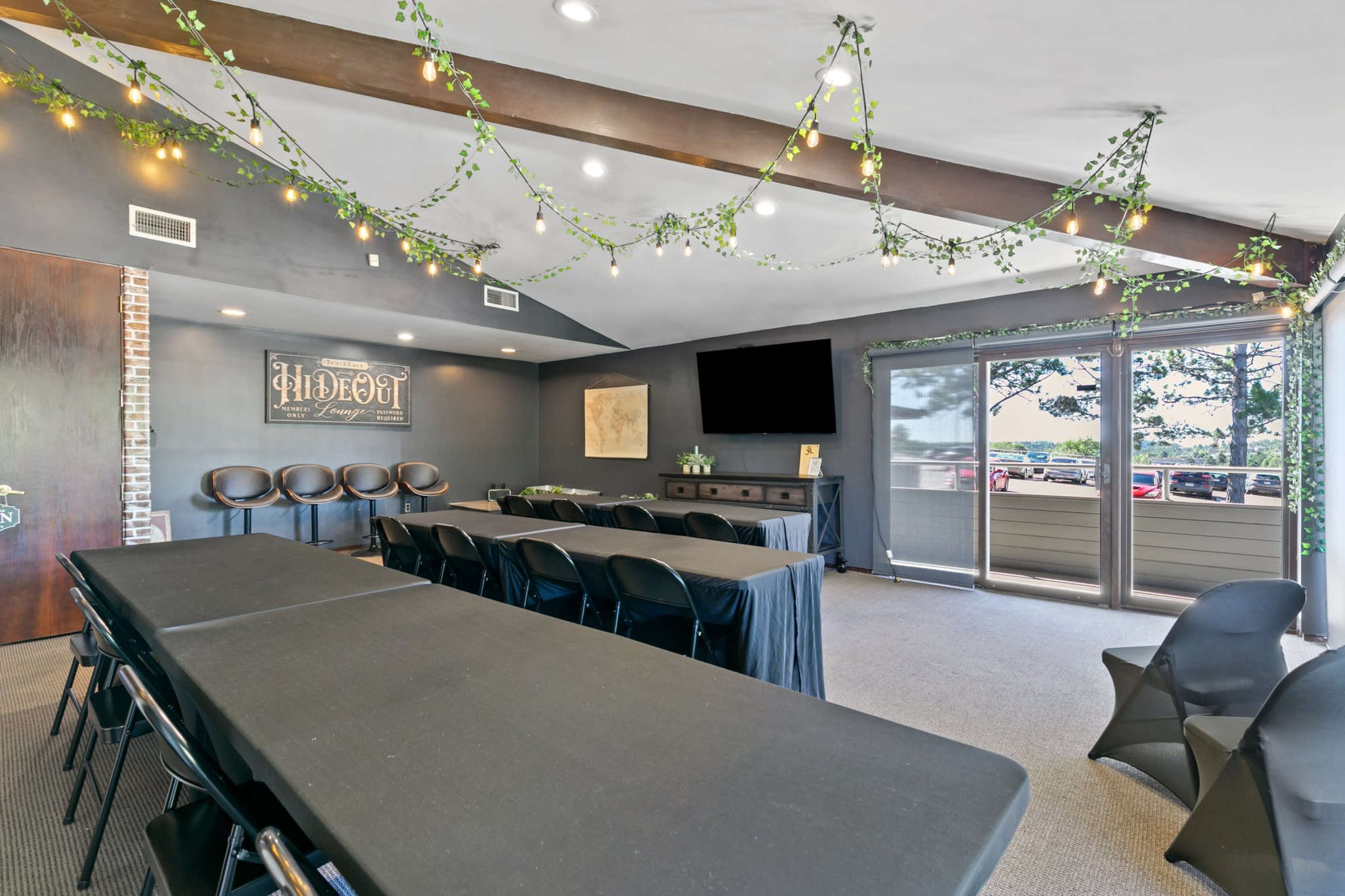 A modern meeting room with black tables and chairs, surrounded by greenery and decorative lights, and featuring a large screen and sliding glass doors that lead to an outdoor view.