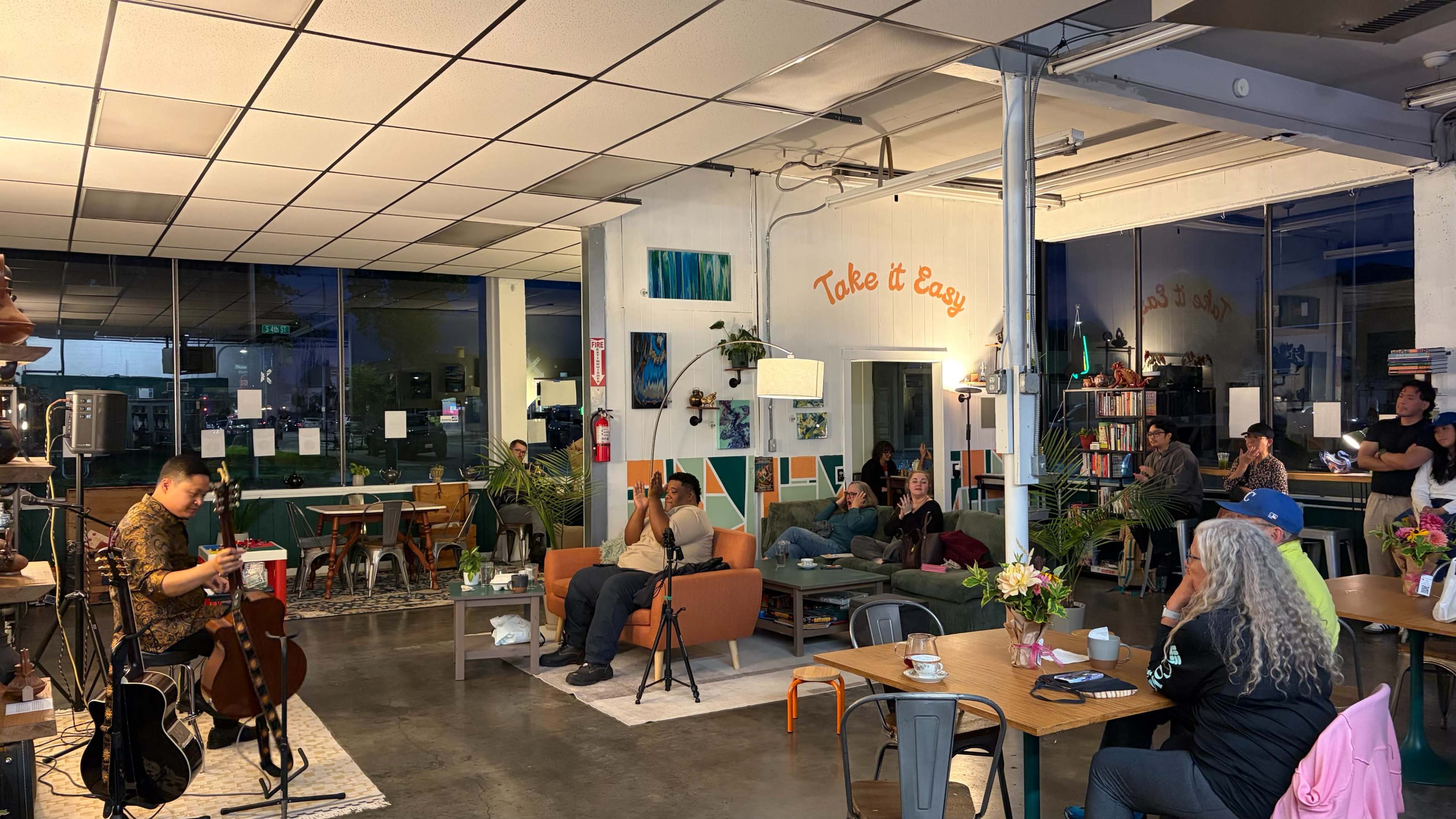 A musician performs on stage in a cozy cafe filled with patrons seated at tables and lounging on sofas.