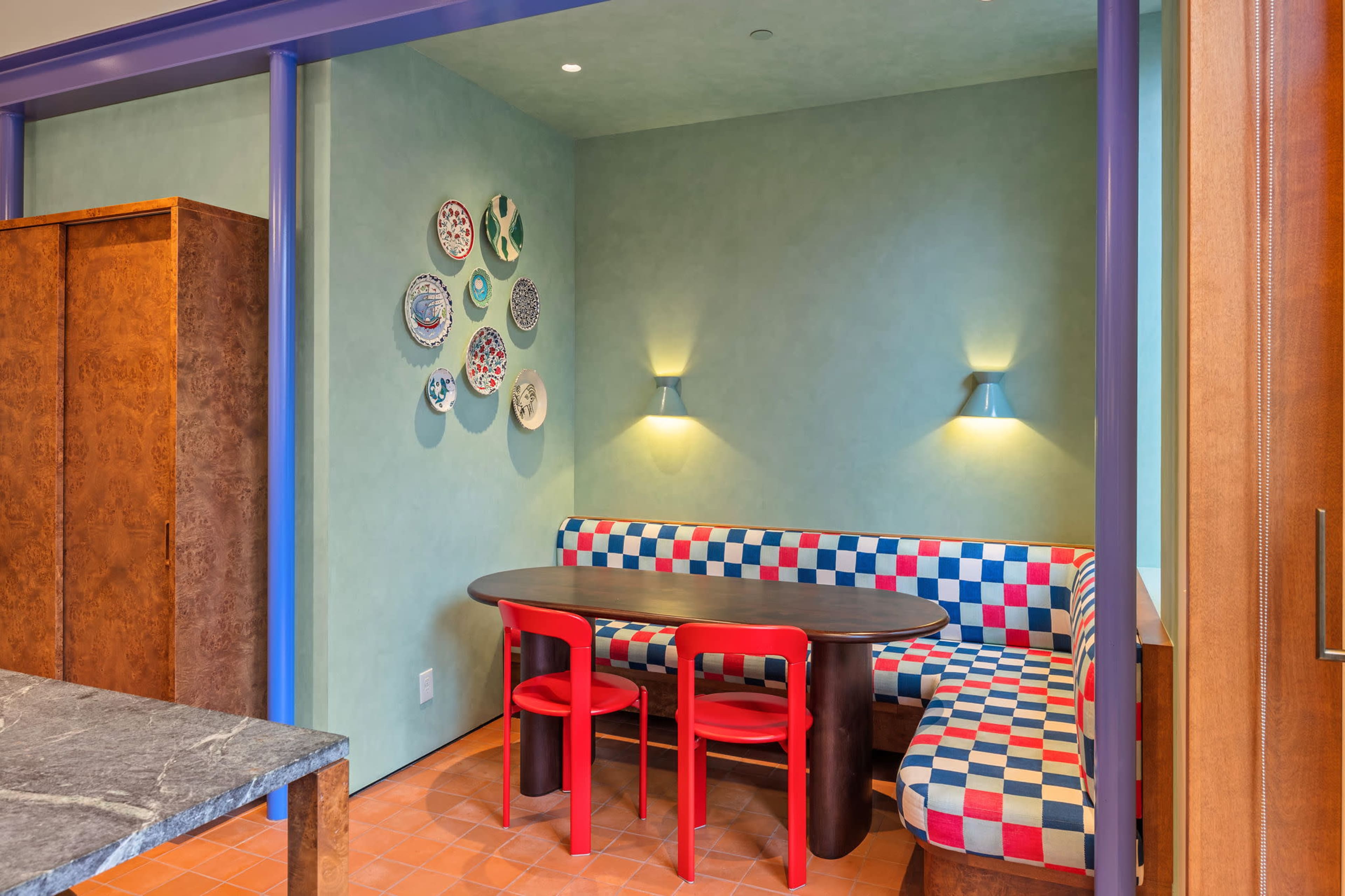 The image shows a small dining area featuring a checkered seating booth with a wooden table and red chairs, alongside decorative plates displayed on the wall.