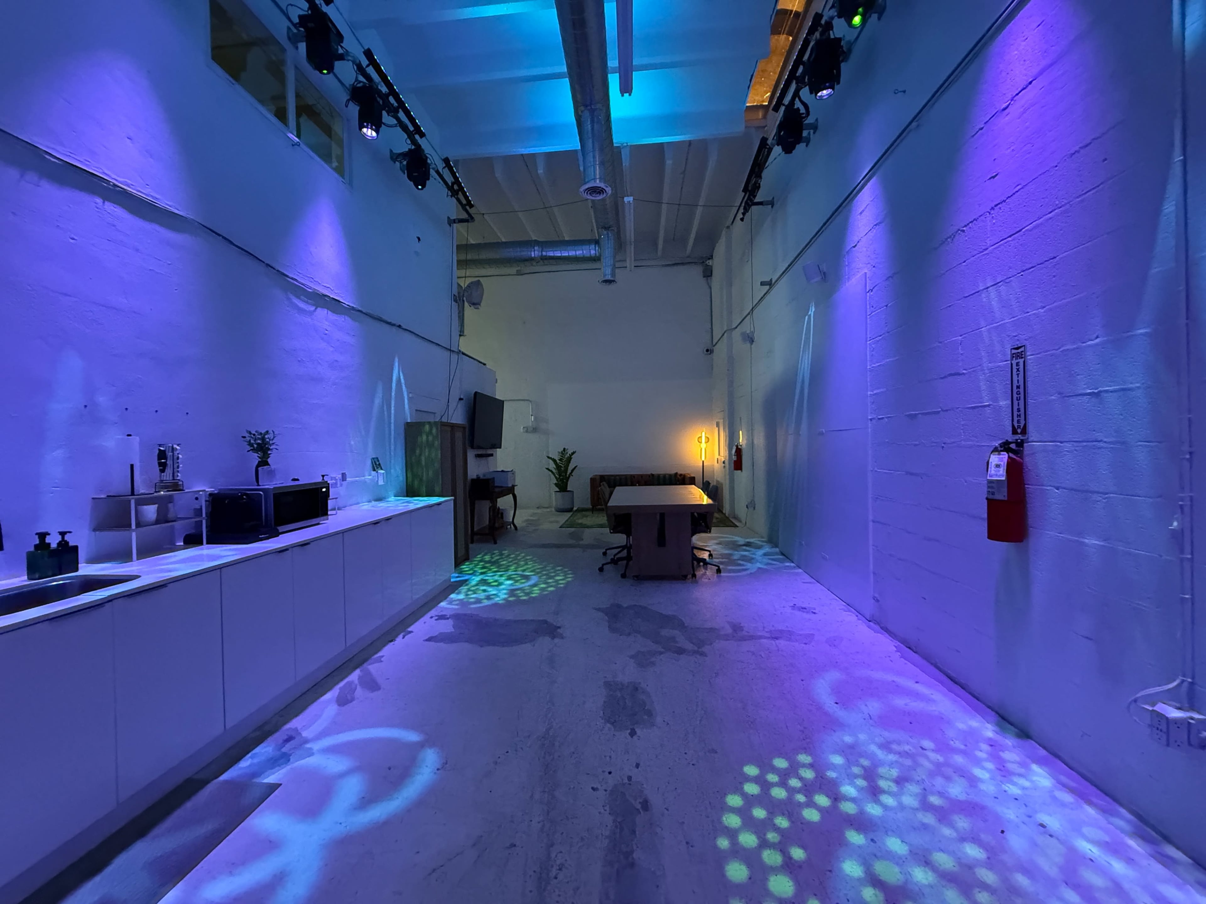 The image shows a spacious, well-lit interior with colorful lights illuminating the walls and floor, featuring a kitchenette along one side and a table in the center.