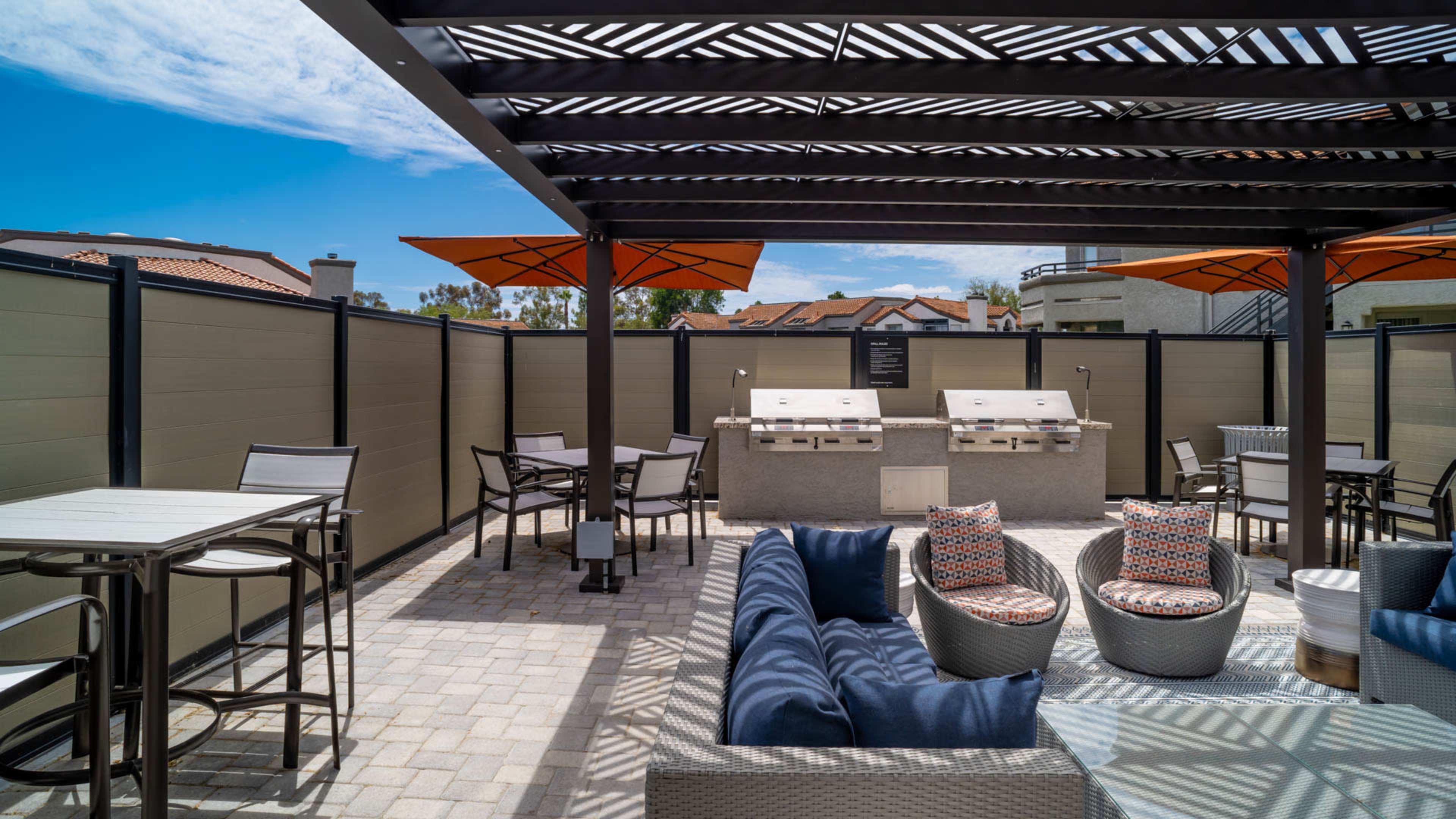 An outdoor grilling area with modern furniture, a stone grill station, and shaded seating under a pergola.