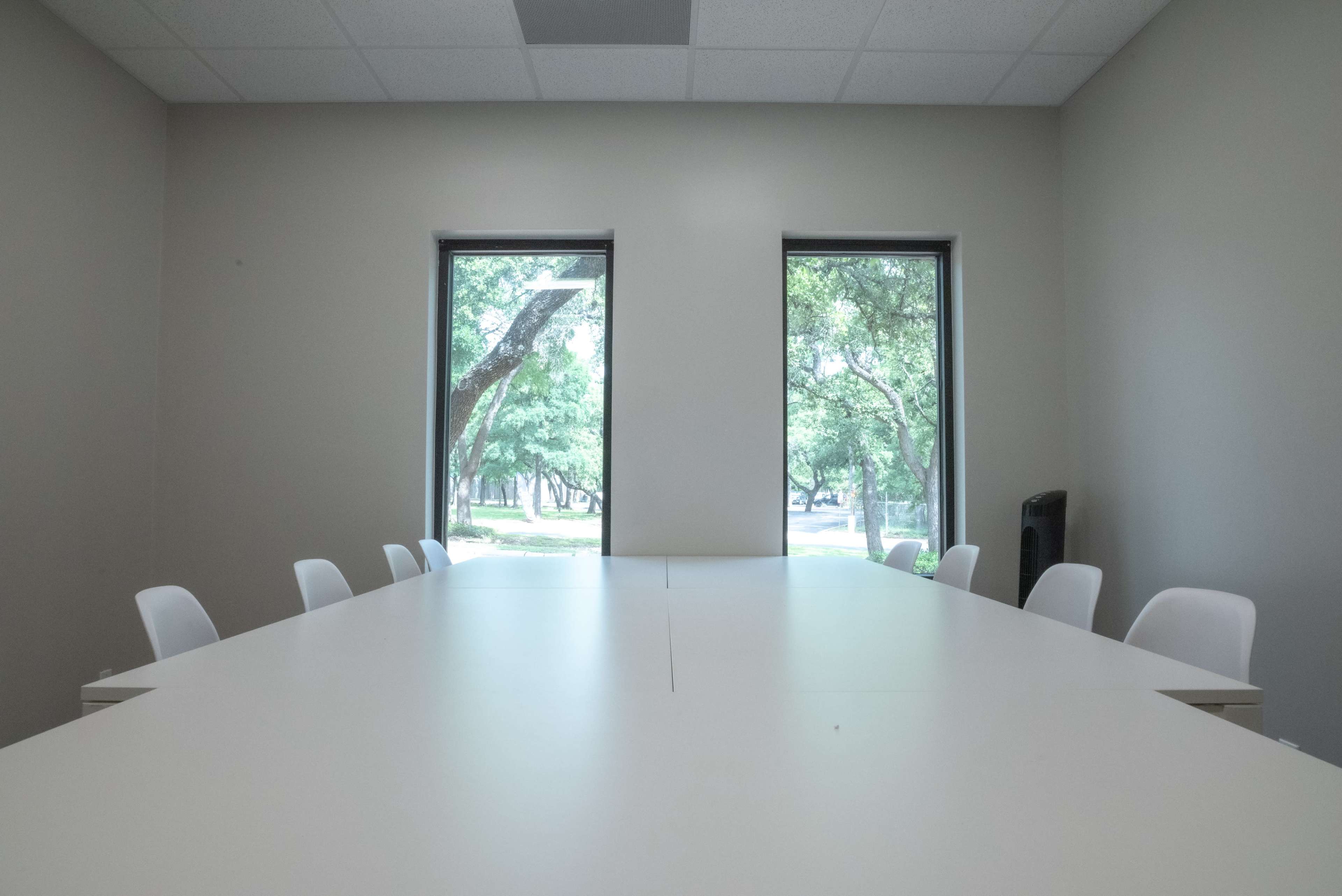 A long white table is set in a meeting room with two windows overlooking trees outside.