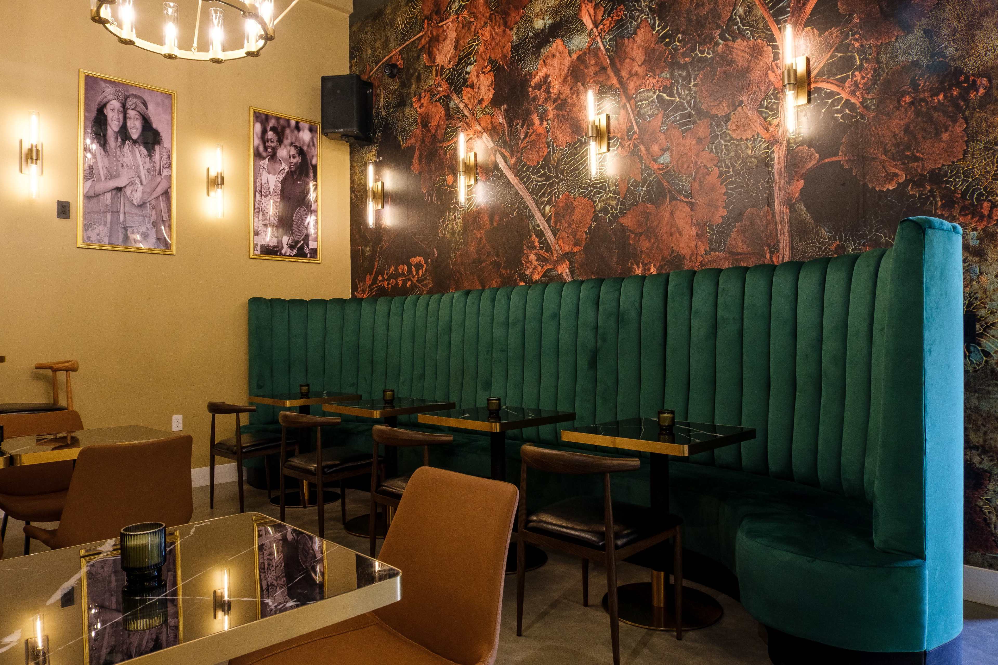 The image shows a modern cafe interior with green velvet seating, gold accents, and floral wallpaper, featuring tables arranged in a cozy setting.