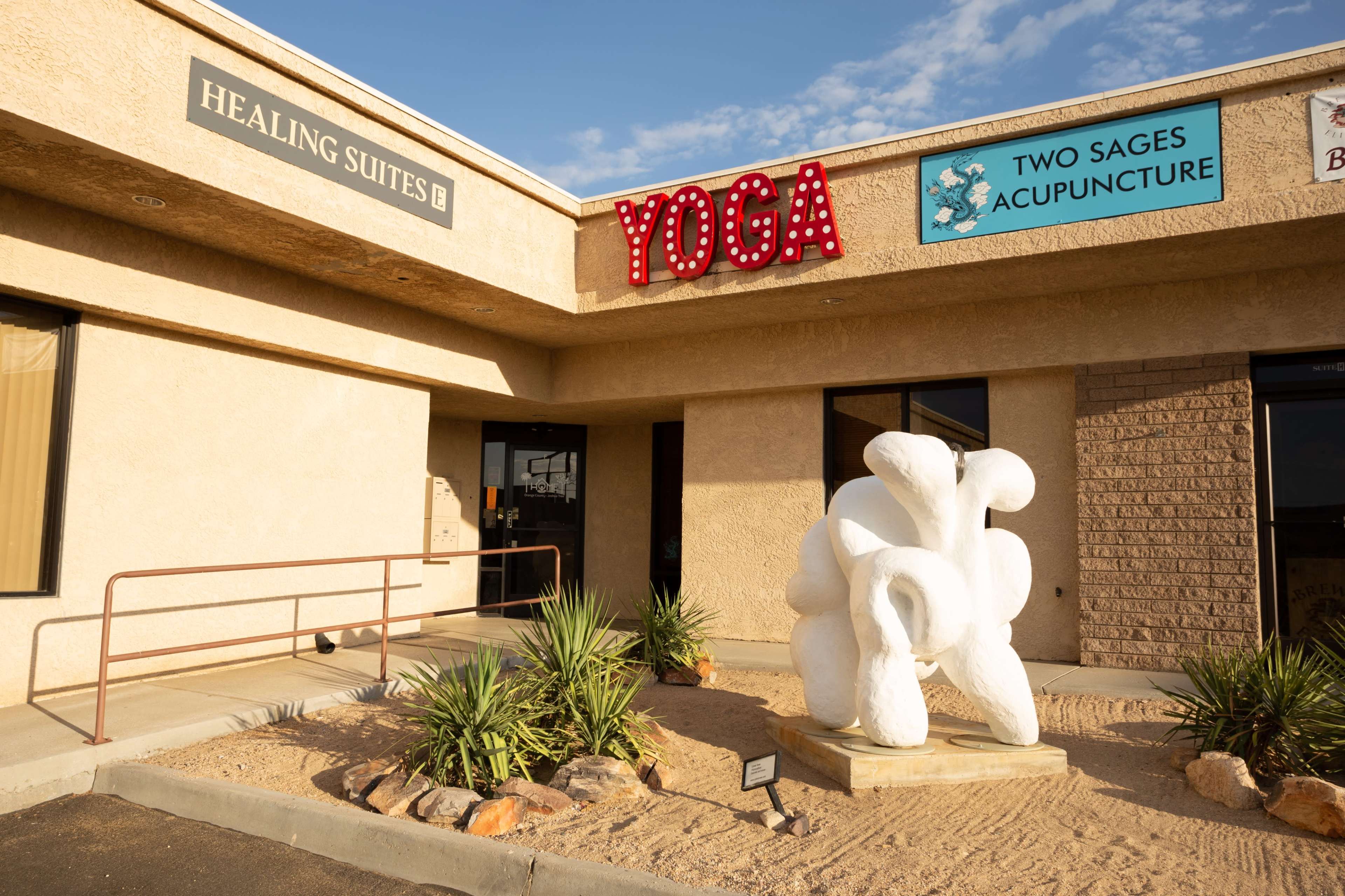 The entrance of a building featuring signs for "Healing Suites," "YOGA," and "Two Sages Acupuncture," along with a white sculpture in front surrounded by desert landscaping.