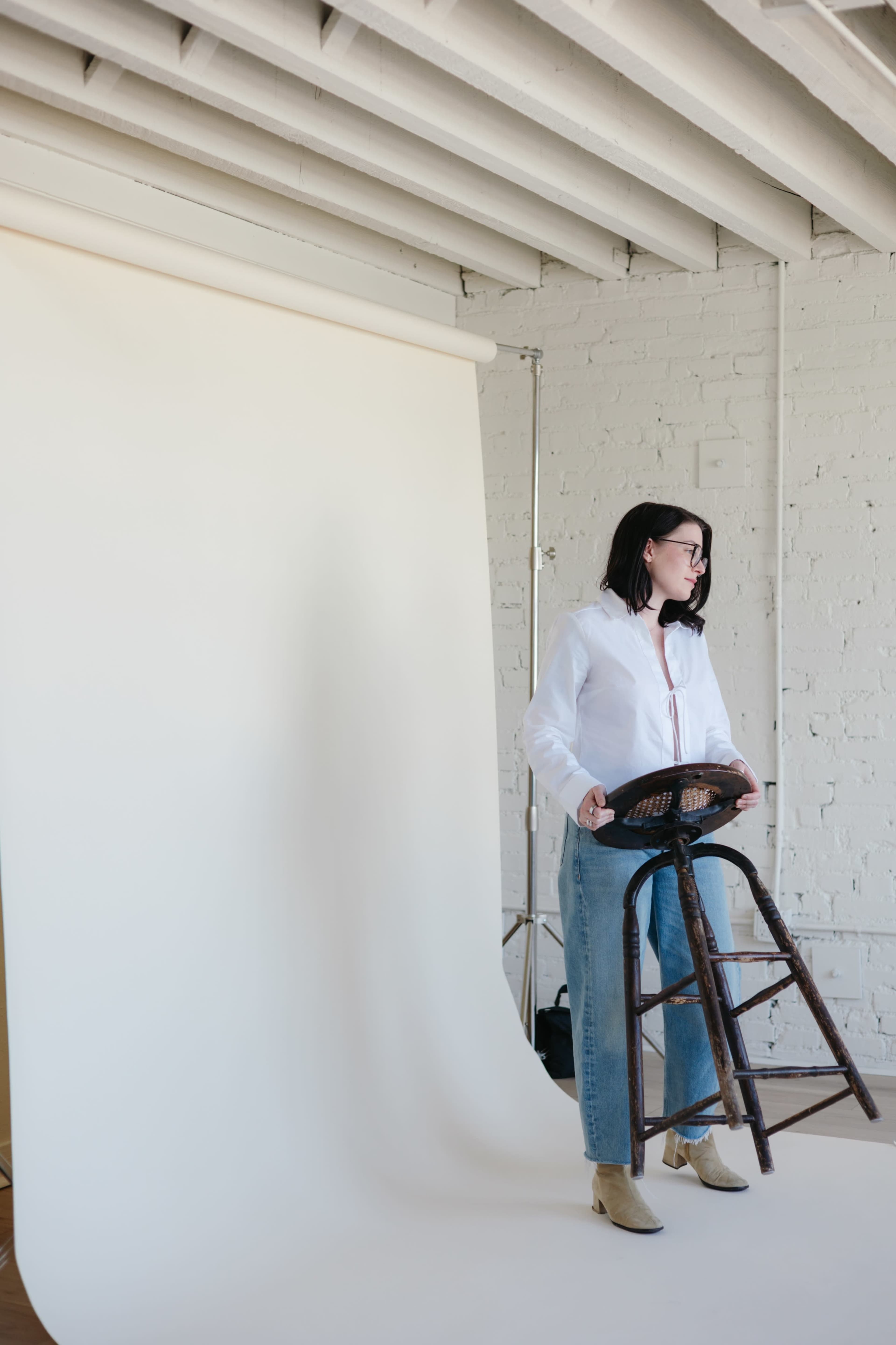 A person stands holding a wooden stool in front of a neutral backdrop in a studio setting.