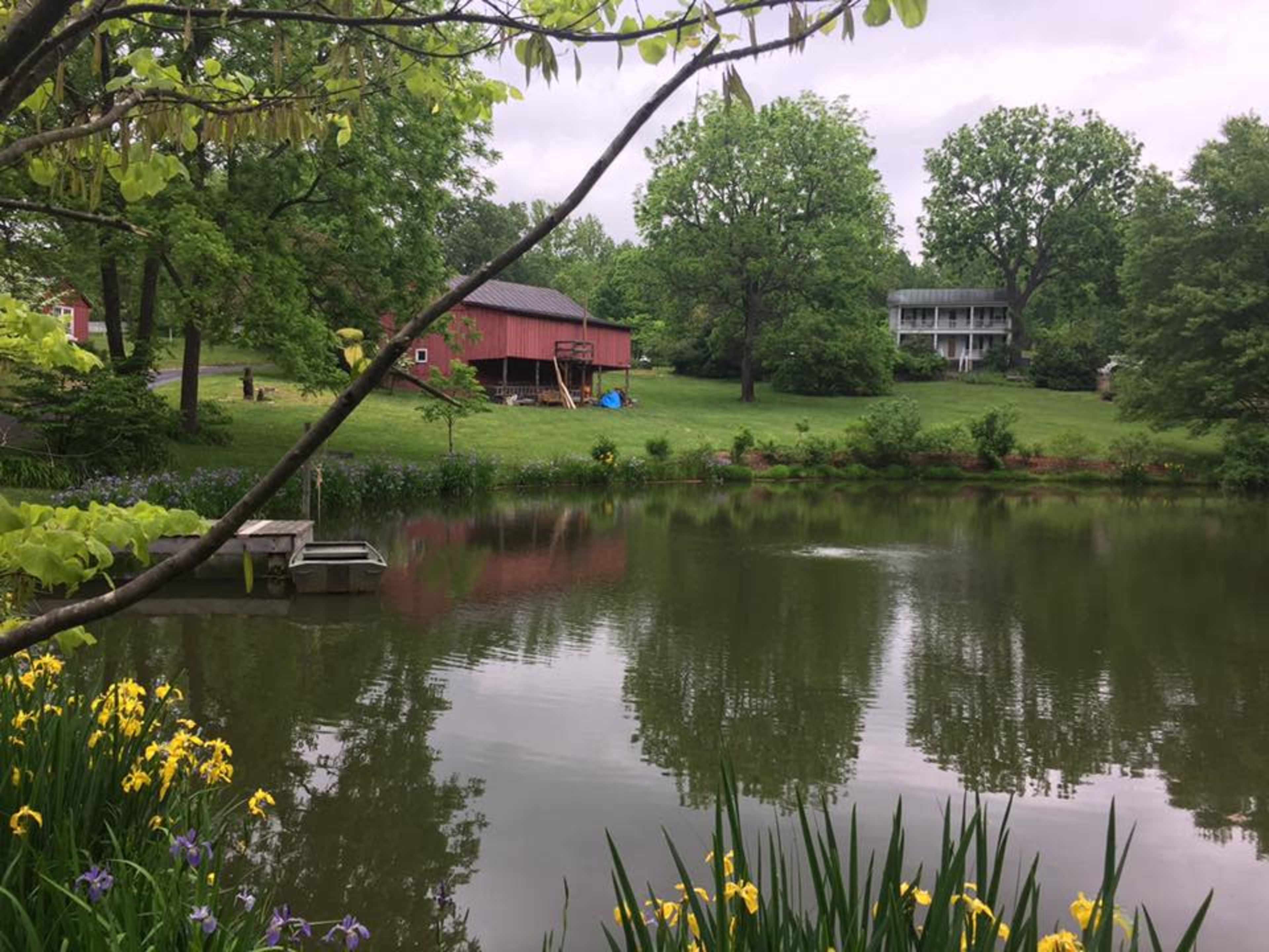 A serene pond reflects the surrounding green trees and a red barn in a rural landscape.