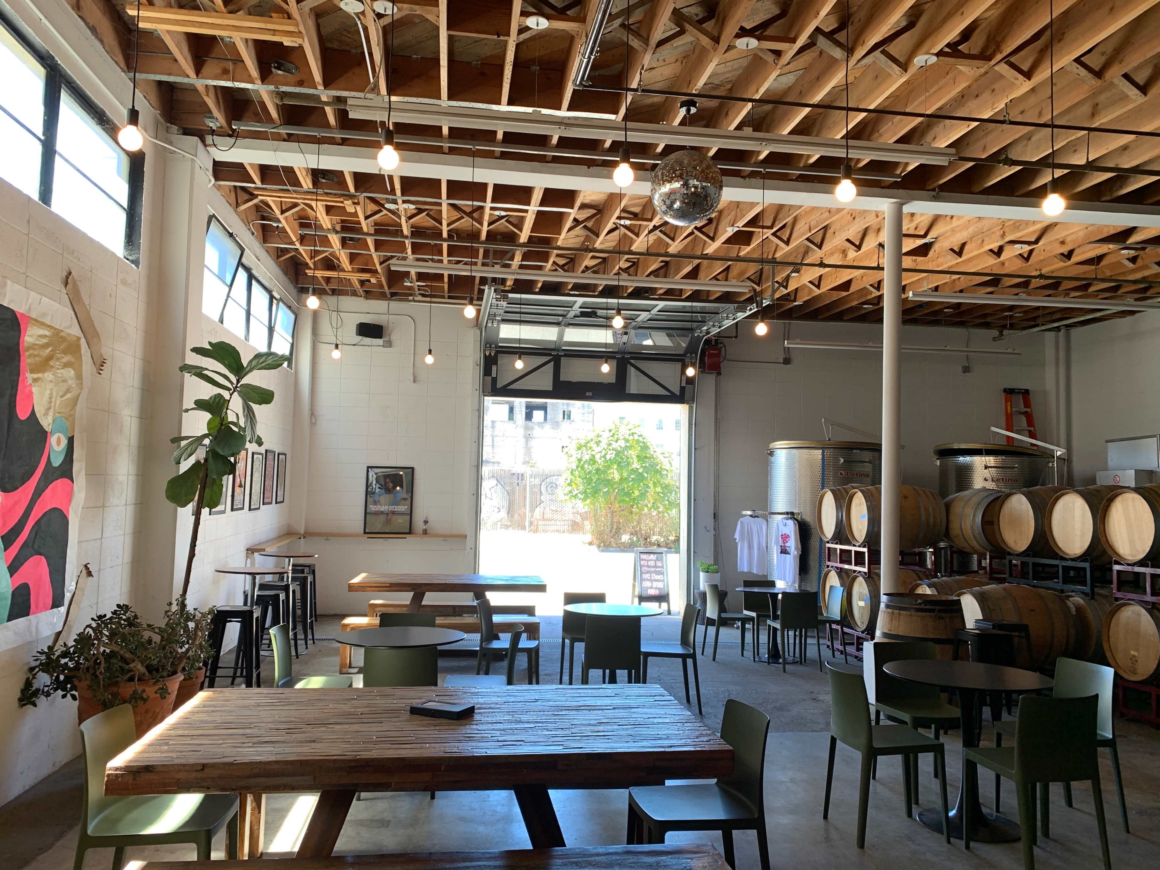 The image shows a spacious, industrial-style interior of a brewery or taproom, featuring wooden tables, a disco ball, and large fermentation tanks in the background.
